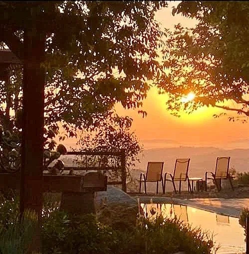 Atardecer con sillones frente a un paisaje con árboles y un cuerpo de agua reflectando el cielo anaranjado.