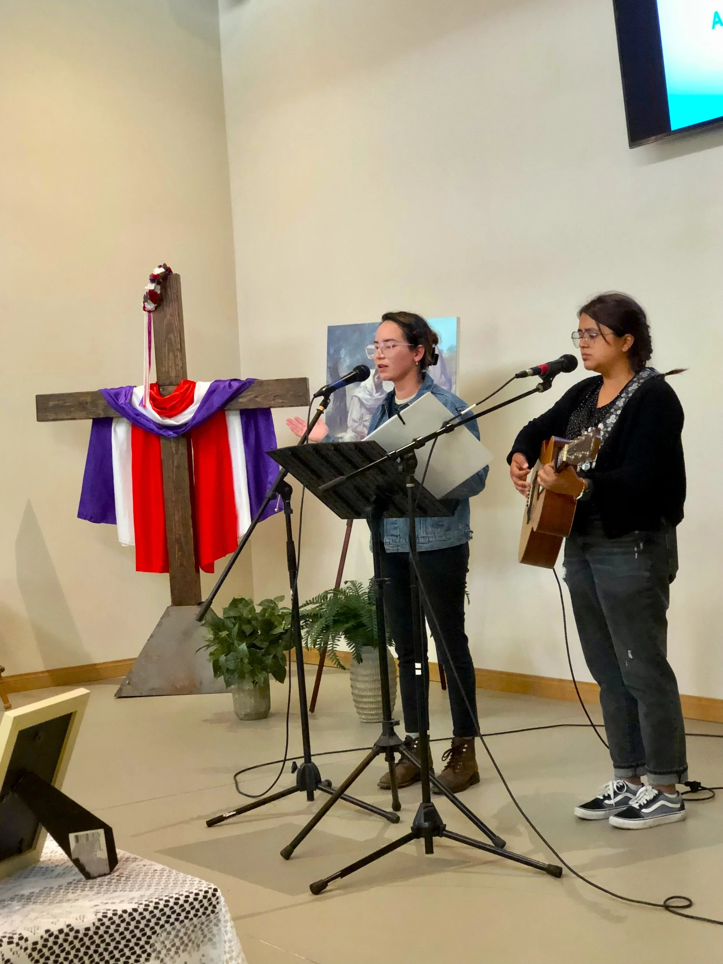 Dos mujeres cantando y tocando guitarra en una iglesia, junto a una cruz decorada con tela tricolor y plantas en un escenario.