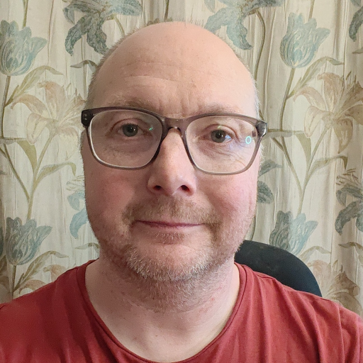 A man with glasses and a short beard wearing a red shirt, seated in front of a floral patterned wallpaper background.