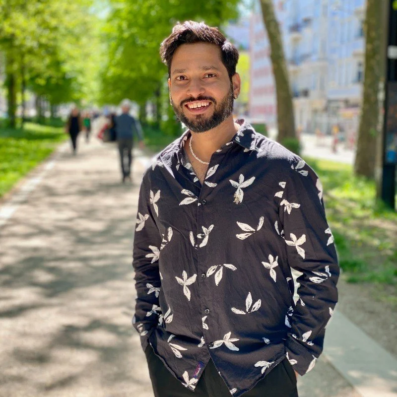 Man with dark hair and beard smiling on a sunny park sidewalk, wearing a black shirt with white leaf patterns, with trees and buildings in the background.