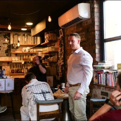 A man in a white shirt standing inside a cozy cafe with brick walls, bookshelves, and warm lighting, with other customers seated at tables.