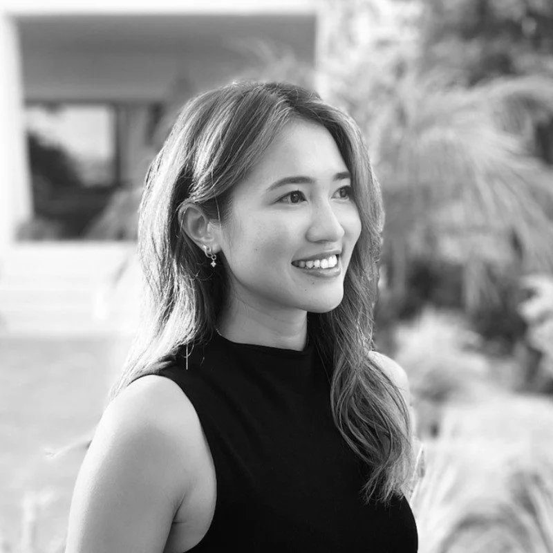 A young woman with light skin and wavy hair smiling outdoors in a sleeveless black top, surrounded by blurred foliage.
