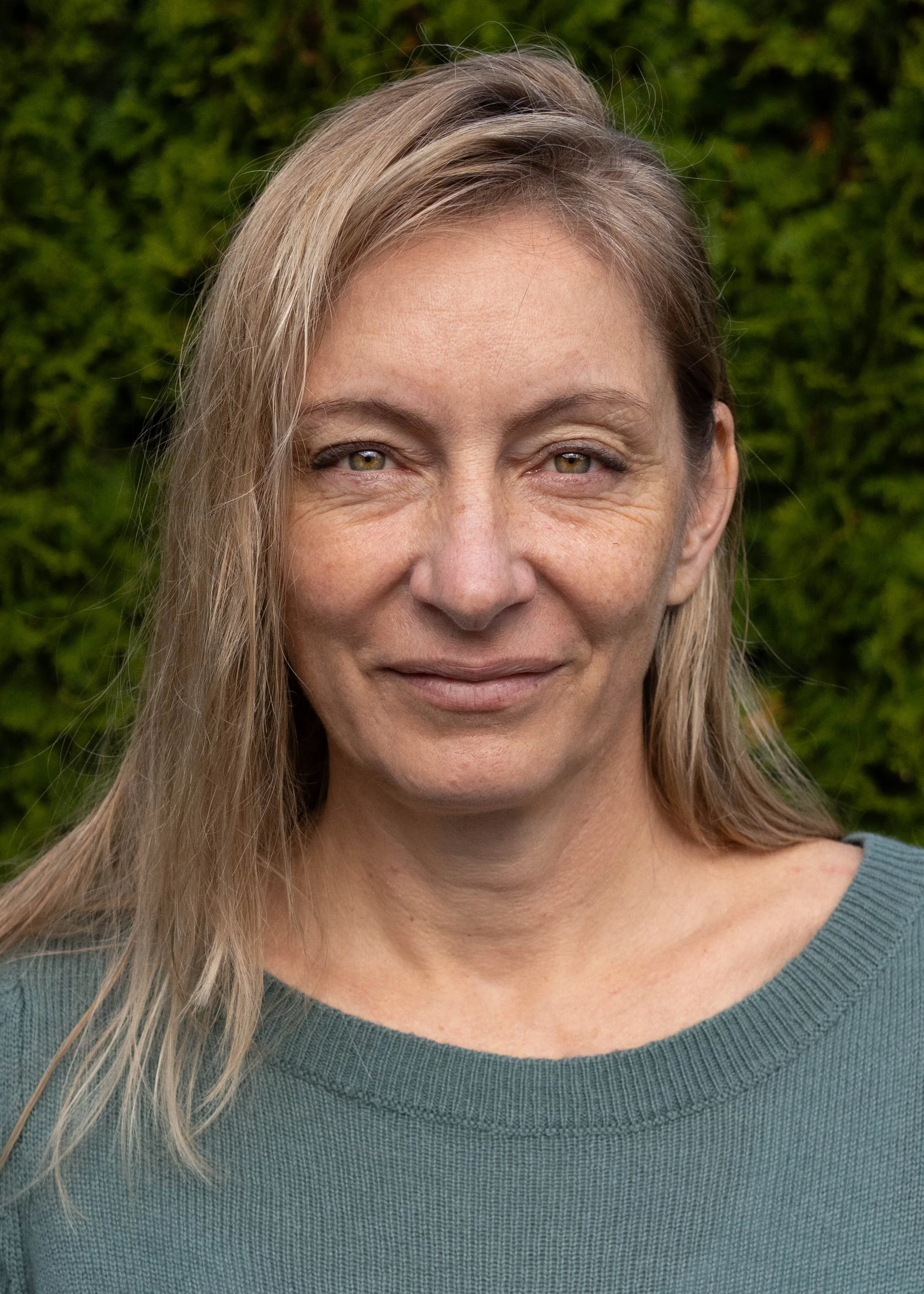 Close-up of a middle-aged woman with blonde hair, wearing a teal sweater, standing outdoors in front of green foliage.