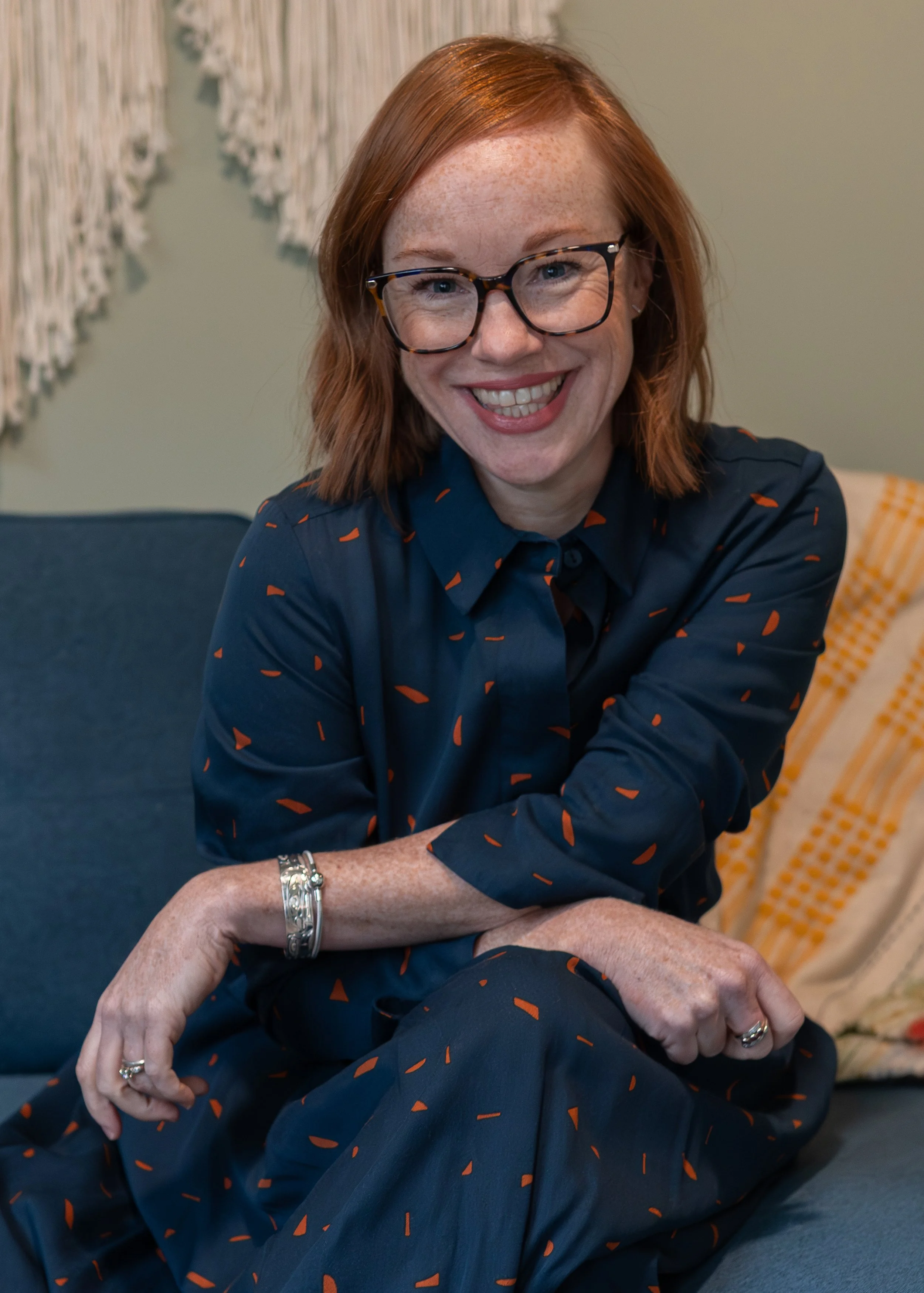 A smiling woman with red hair, glasses, and freckles, sitting on a blue couch, wearing a dark blue dress with orange patterns, accessorized with jewelry.