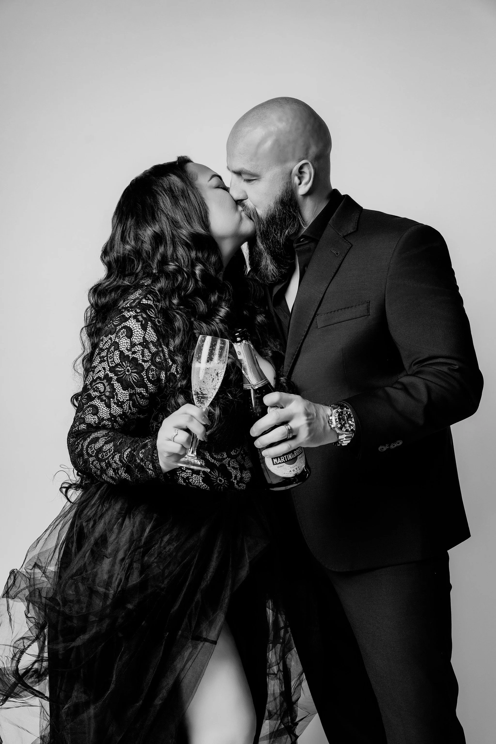 A black and white photo of a couple kissing, holding champagne glasses and a bottle, dressed in formal attire.