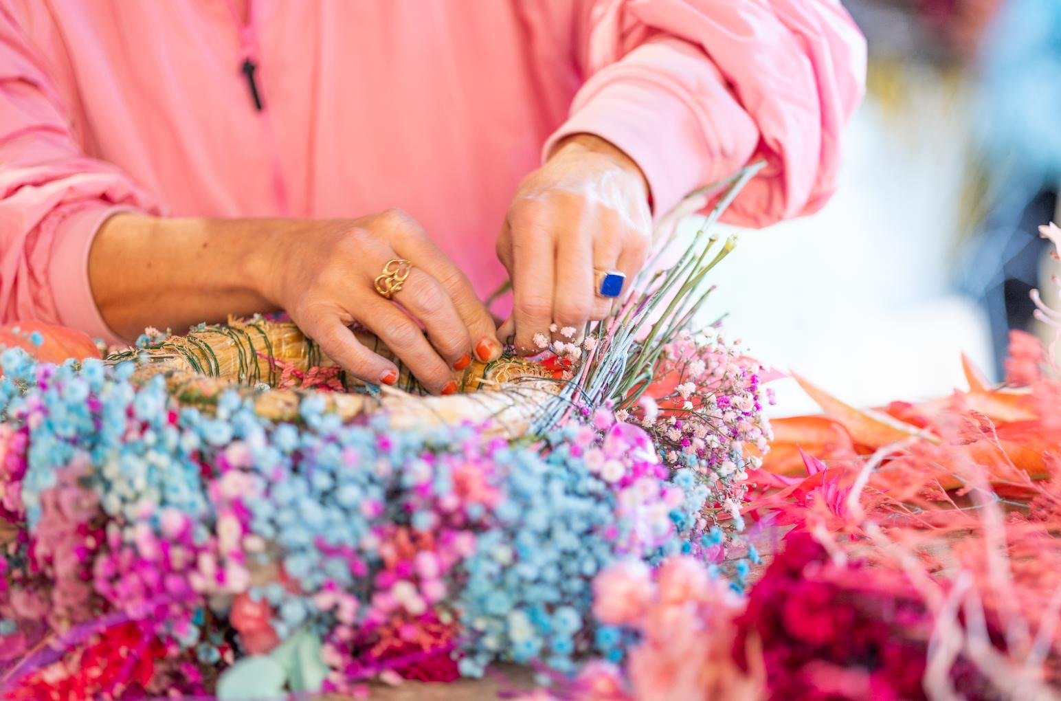 Person bindet bunte Blumen an einen Korb, trägt einen pinken Jacke, trägt Ringe an den Fingern, mit farblich passenden Blumen.