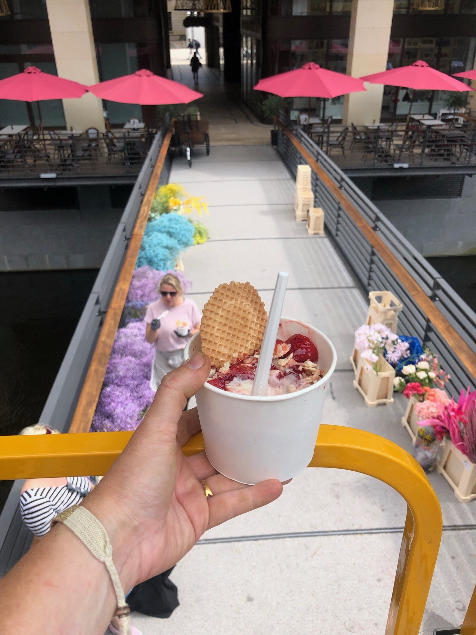 Eine Hand hält ein Becher Eis mit Früchten und Waffel in einer oberen Perspektive, im Hintergrund ein Balkon mit roten Sonnenschirmen, Blumen und einer Person.