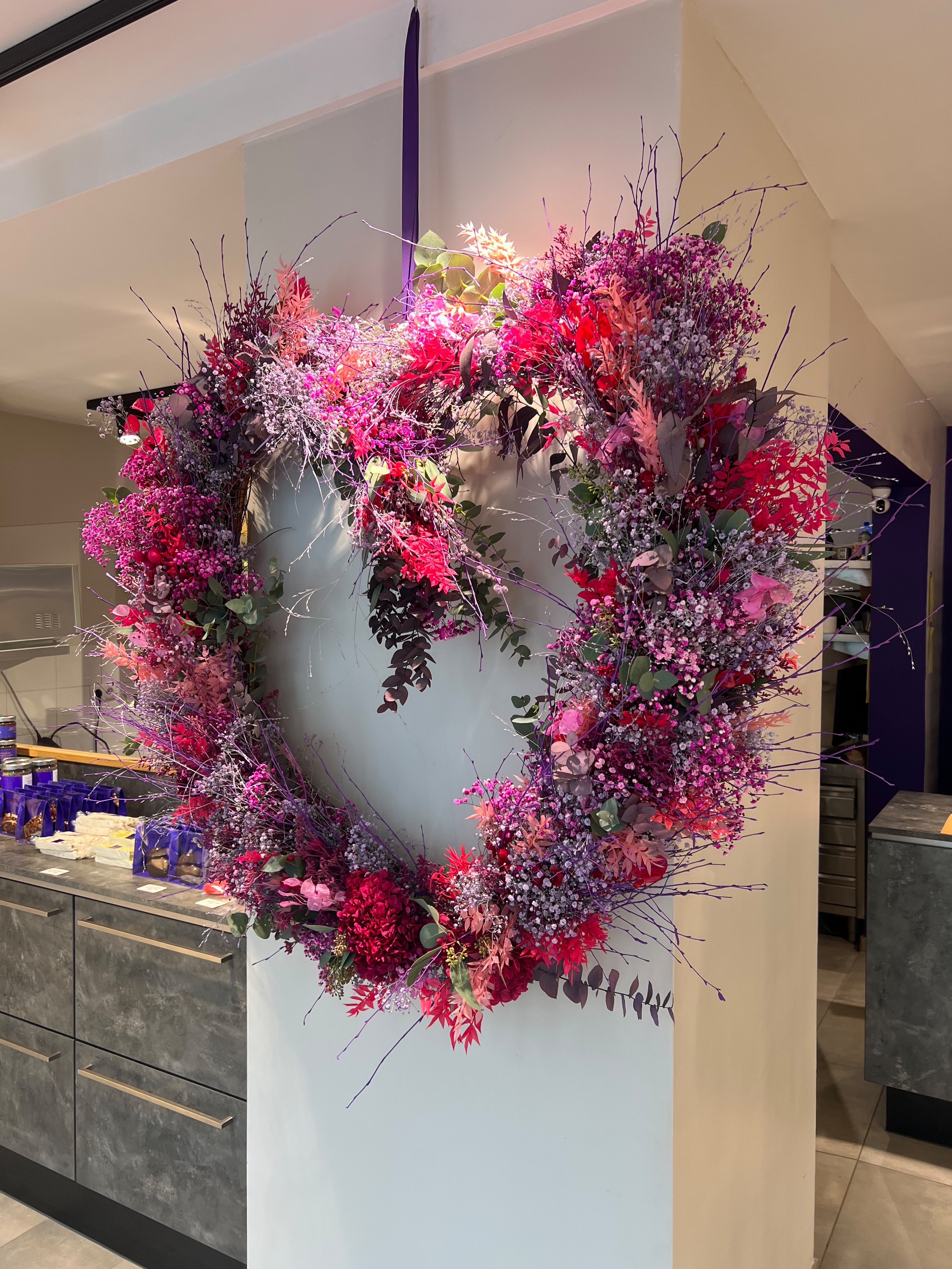 Blumenkranz aus roten, pinken, lila und weißen Blumen und Zweigen, hängen an einer Wand in einem Raum.