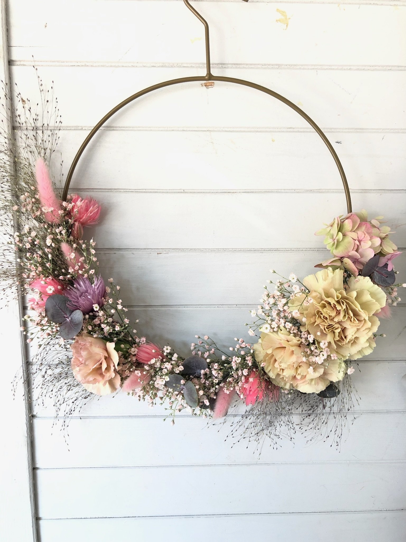 Blumekranz aus rosa, lavendel, cremefarbenen und grünen Blumen mit grauen Blättern und Federn, dekorativ an einer Wand aufgehängt.
