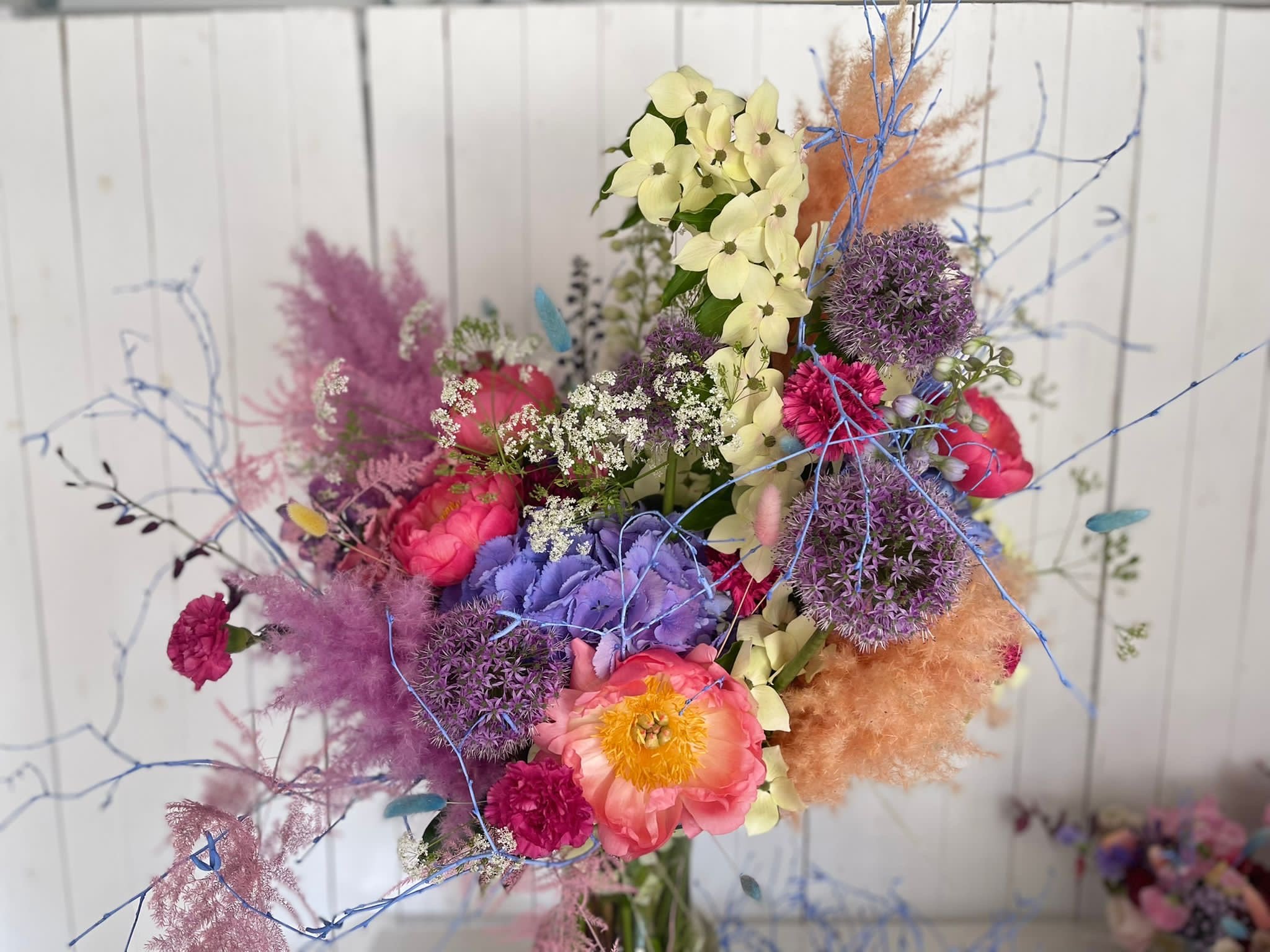 Bunter Blumenstrauß mit verschiedenen Blumen wie Pfingstrosen, Hortensien und Allium in einer Vase, vor einem weißen Holzwand