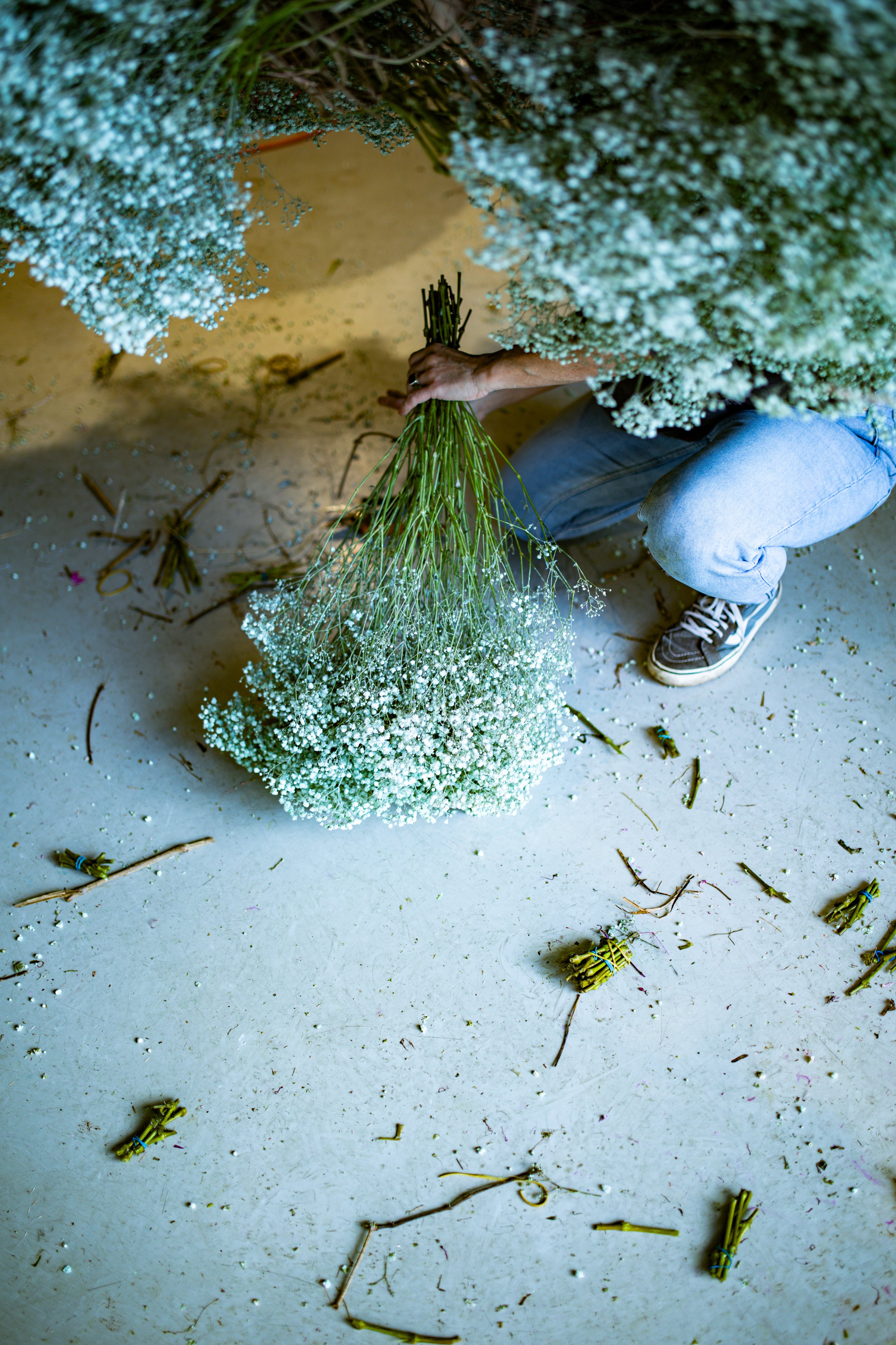 Person beim Blumenbinden von weißen Schleierkraut in einem Raum mit verstreutem Tüll und Blumenstielen auf dem Boden.
