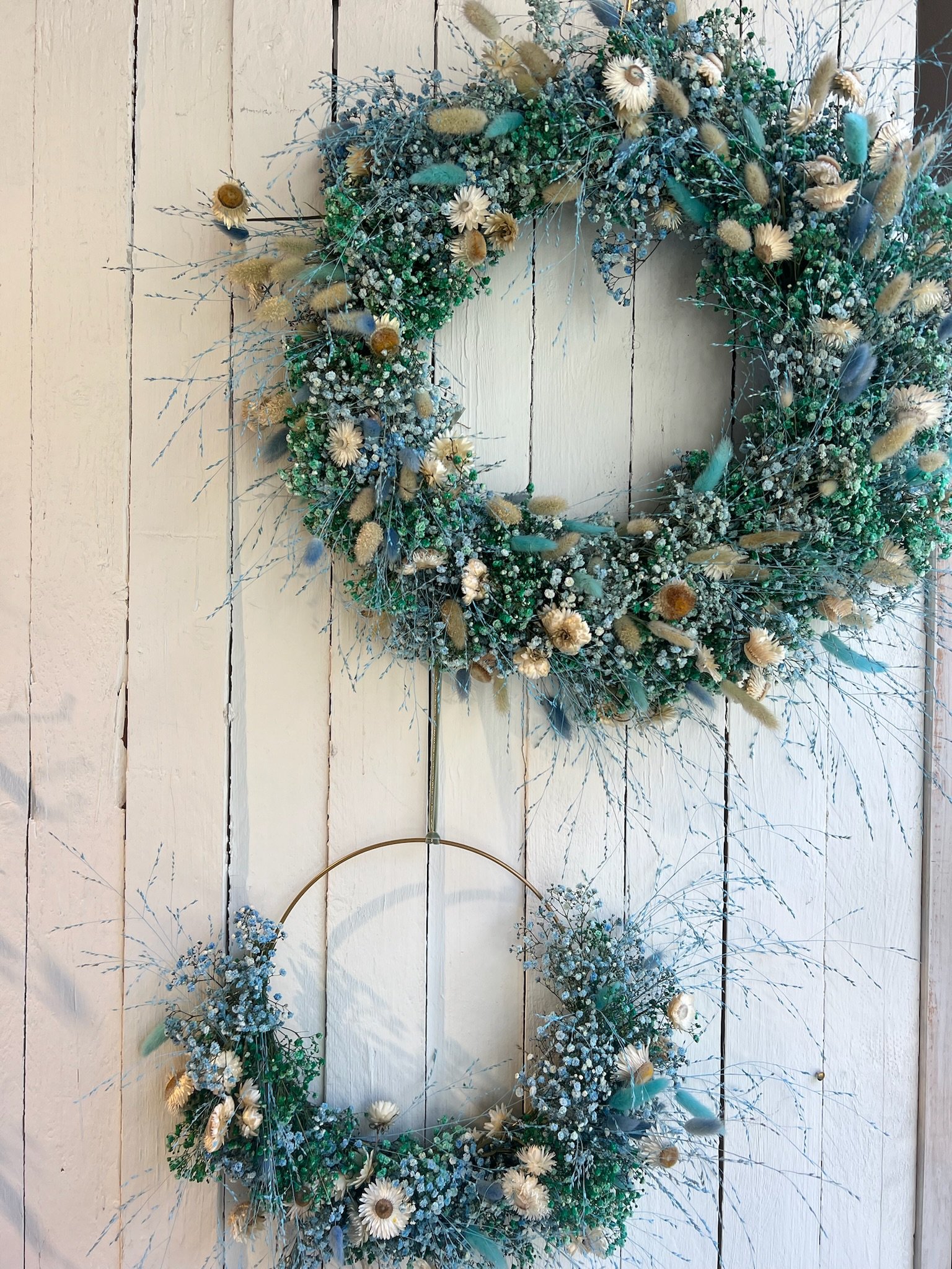 Zwei prächtige Blumenkränze aus getrockneten Blumen, darunter weiße Blüten, blaue Feder und getrocknetes Gras, hängen an einer weißen Holzwand.