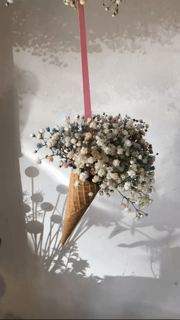 Ein dekoratives Wandbild mit einer Eistüte aus Blumen, die an einer pinken Schleife hängt, mit Schatten an der Wand