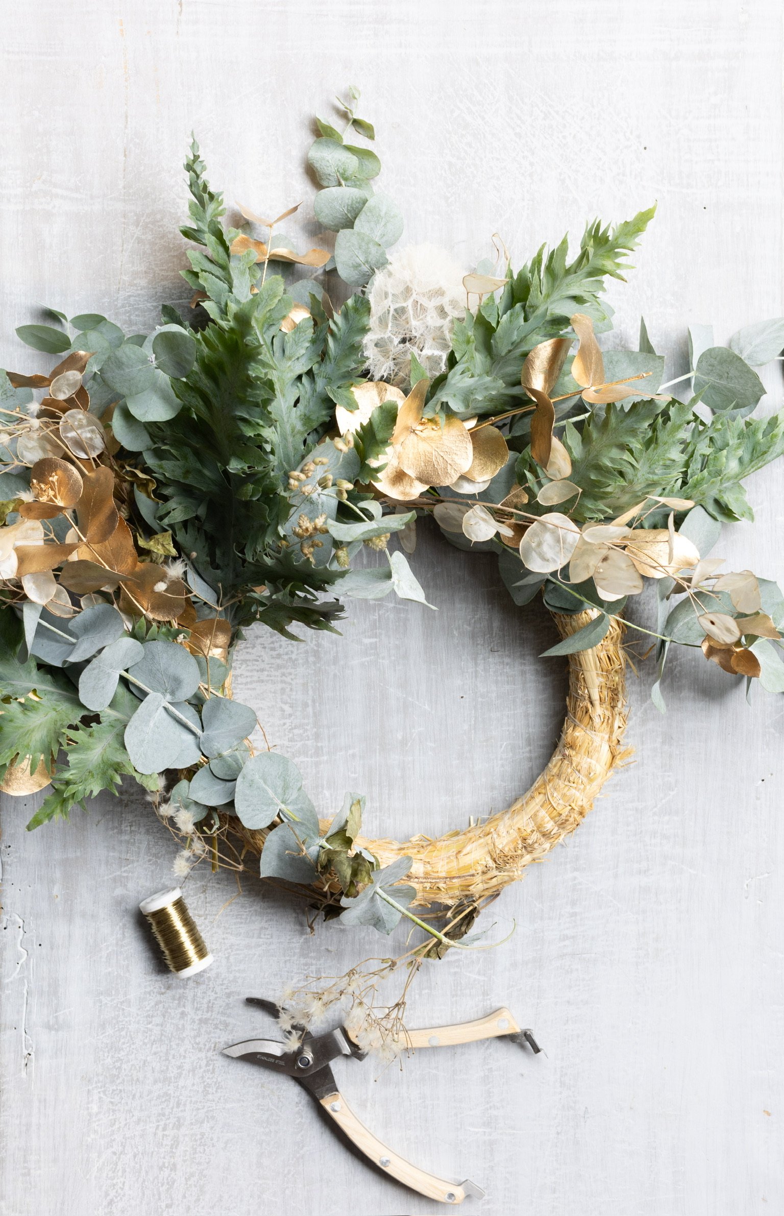 Kränzchen aus Eukalyptus und anderen grünen Blumen mit goldenem Dekor, Werkzeug und Faden auf grauem Untergrund.