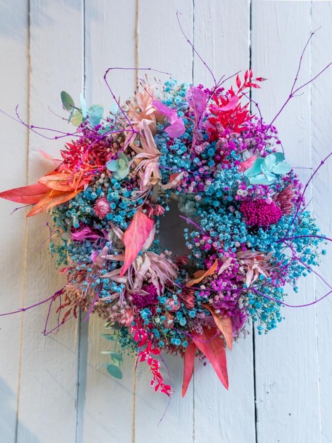 Bunter Blumenkranz mit blauen, pinken und orangefarbenen Blüten an weißem Holz.
