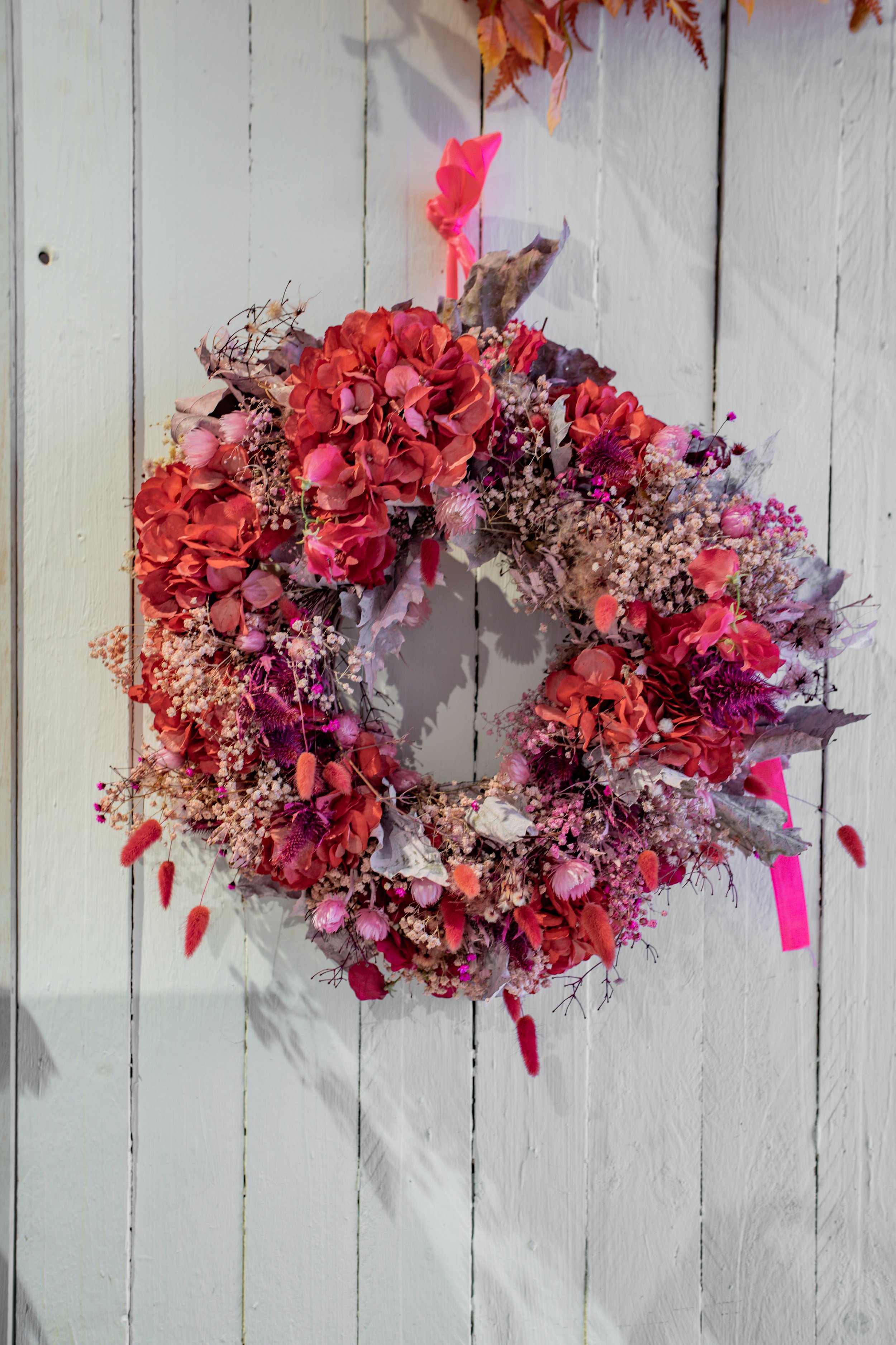 Ein rosa Blütenkranz mit roten, rosa und lilafarbenen Blumen, aufgehängt an einer weißen Holzwand.