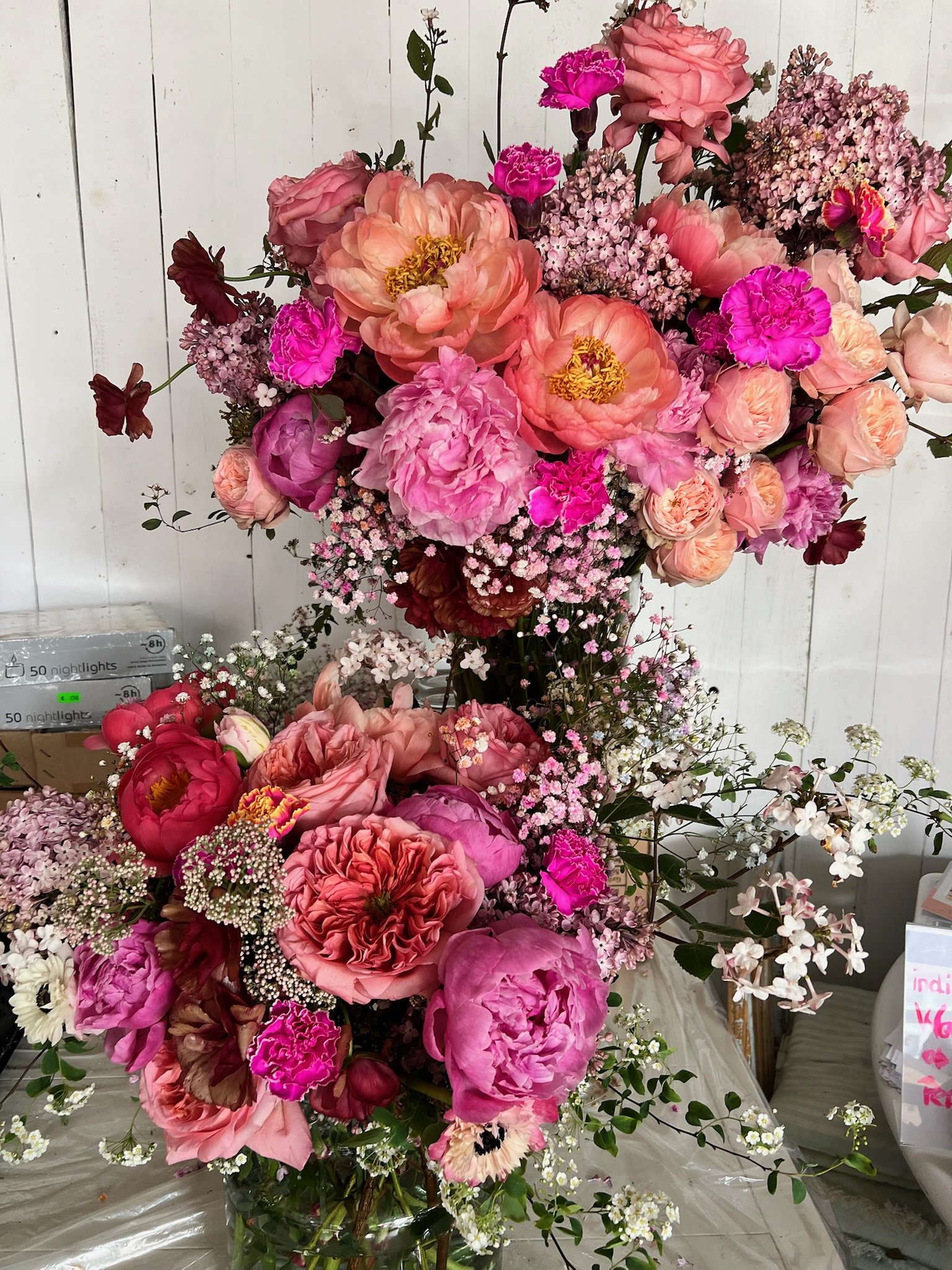 Ein bunter Blumenstrauß mit verschiedenen pinken, roten und peachfarbenen Blumen, darunter Pfingstrosen, Rosen und Flieder, steht in einer Glaskaraffe vor einer weißen Holzwand.