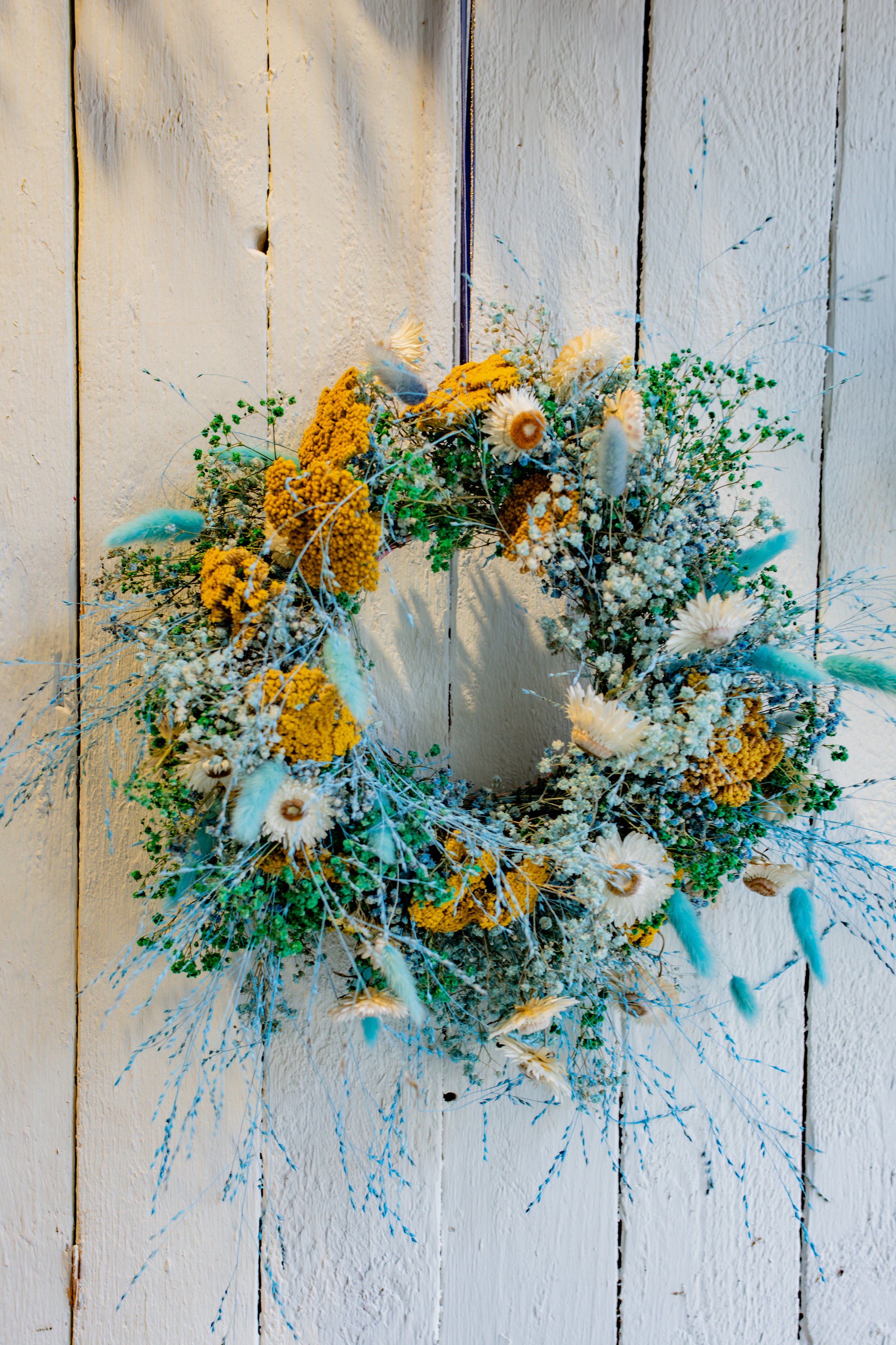 Blumenkranz aus gelben, weißen und blauen Blumen, an einer weißen Holzwand aufgehängt.