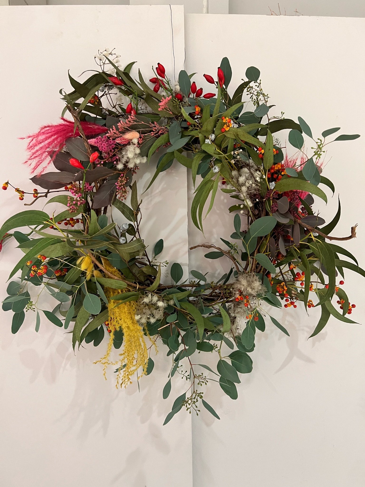 Ein Blumenkranz aus verschiedenen Grünpflanzen und bunten Blumen, einschließlich roter Beeren, gelber und pinker Blüten, an einer weißen Wand.