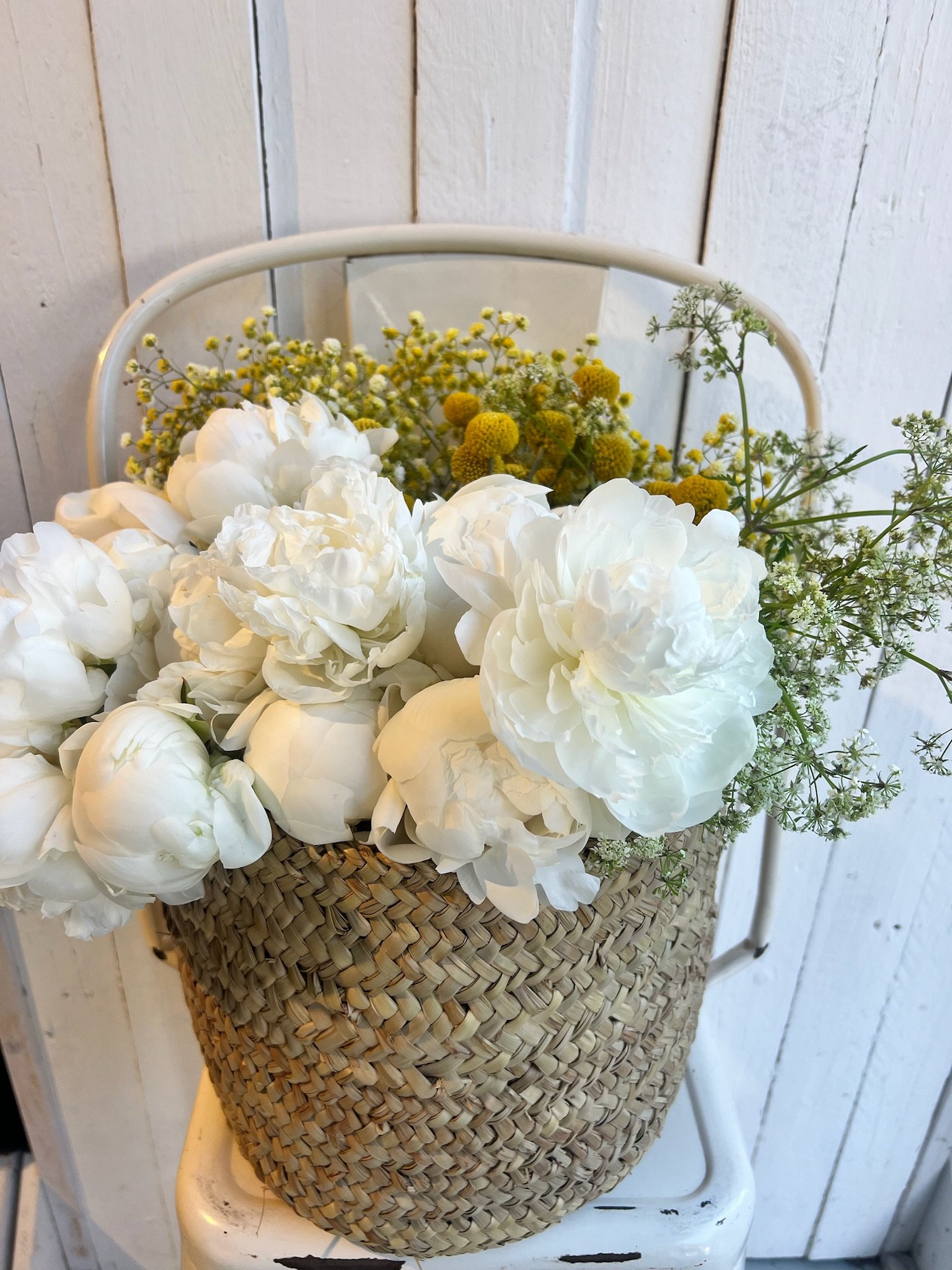 Ein Korb mit weißen Blumen, umgeben von kleinen gelben und weißen Blüten, auf einem weißen Möbelstück gegen eine Holzwand.