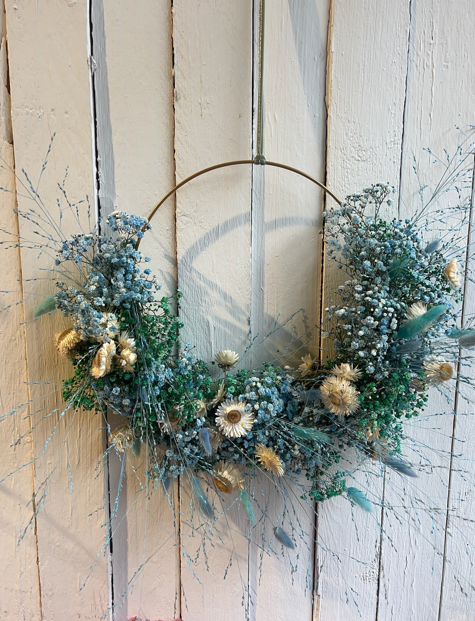 Blumenkranz aus weißen und blauen Blumen, an einem goldenen Ring an einer weißen Holzwand.