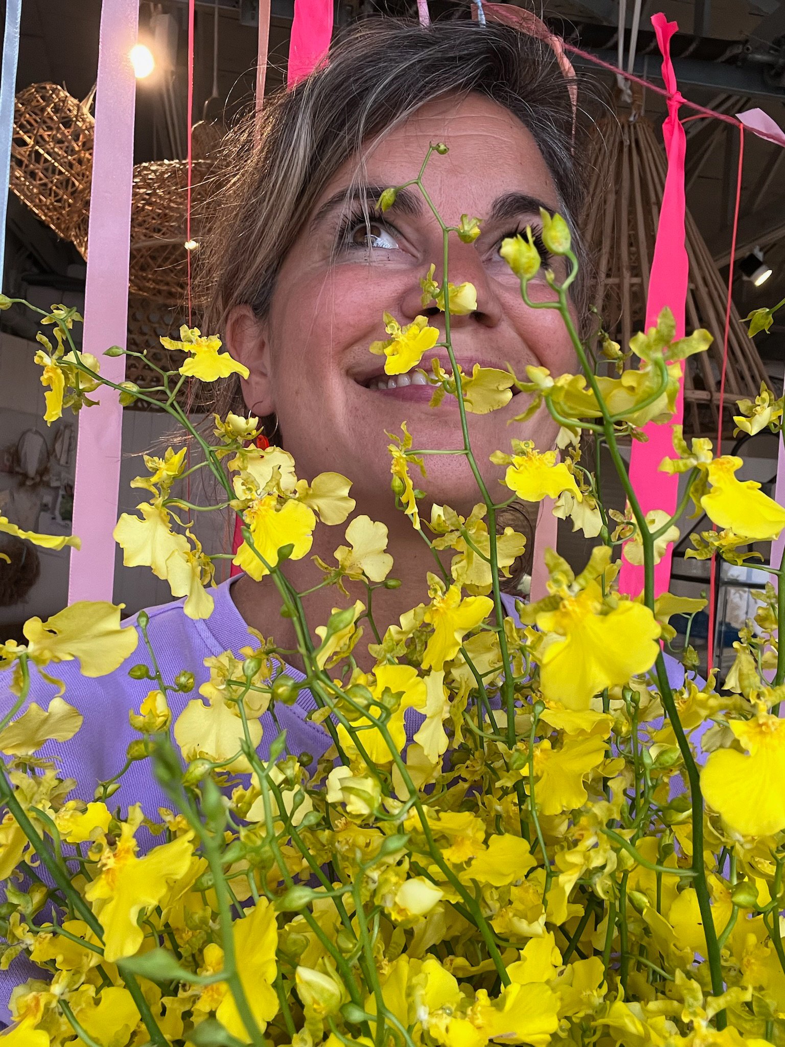Frau hinter einer Vielzahl gelber Blumen, lachend, in einem Raum mit Dekor und Pinken Bändern.