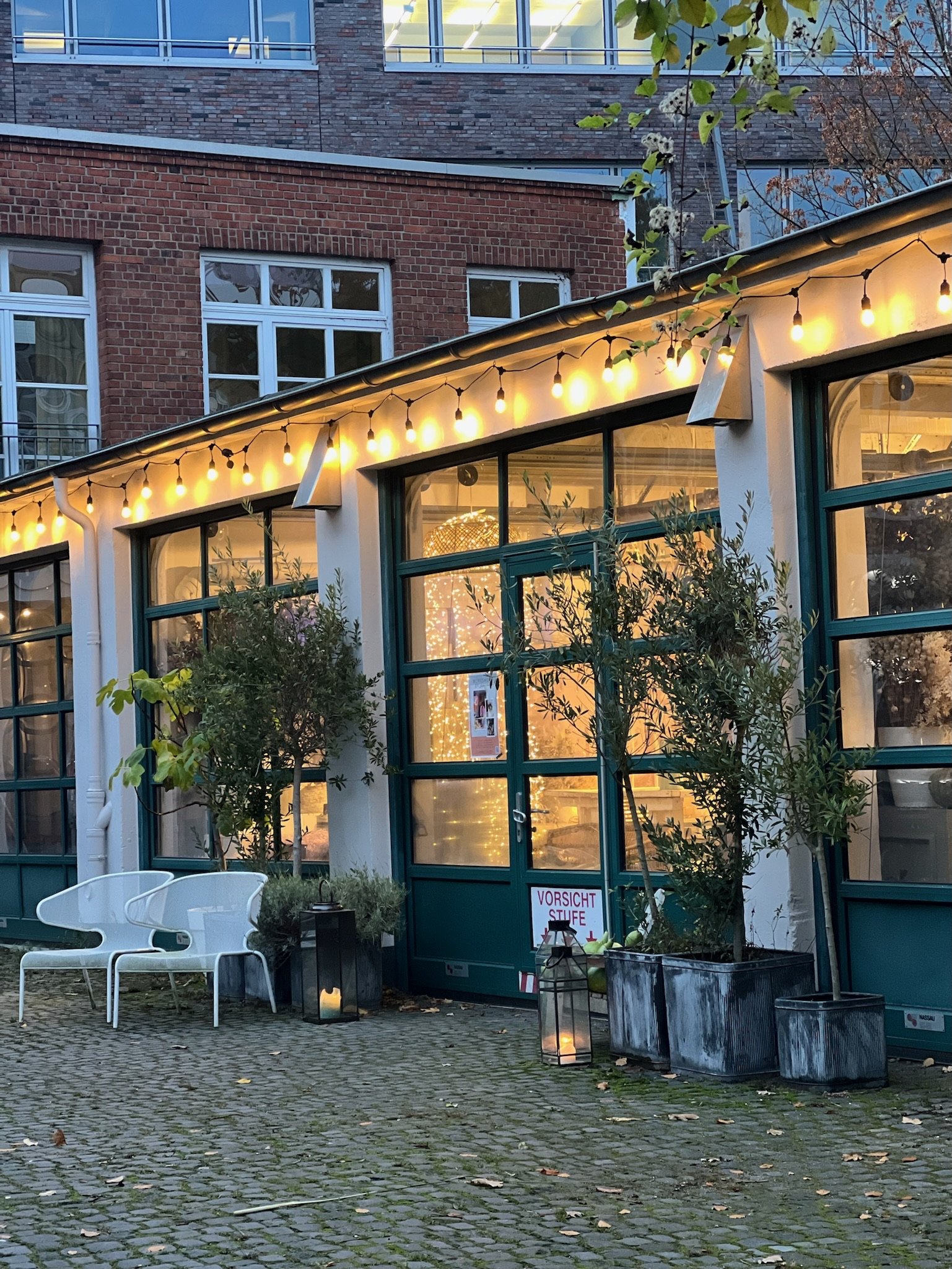 Außenbereich eines Restaurants oder Cafés mit warmem Inneneinblick, blau gerahmte Fenster, Lichterkette unter dem Dach, zwei weiße Gartenstühle, Pflanzen in großen Kästen, Laternen mit Kerzen, cobbelsteinerter Boden.