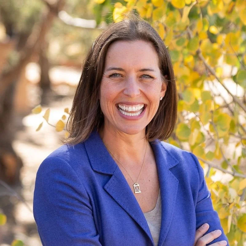 A woman smiling with arms crossed outdoors, wearing a blue blazer and a necklace, with yellow and green leaves in the background.