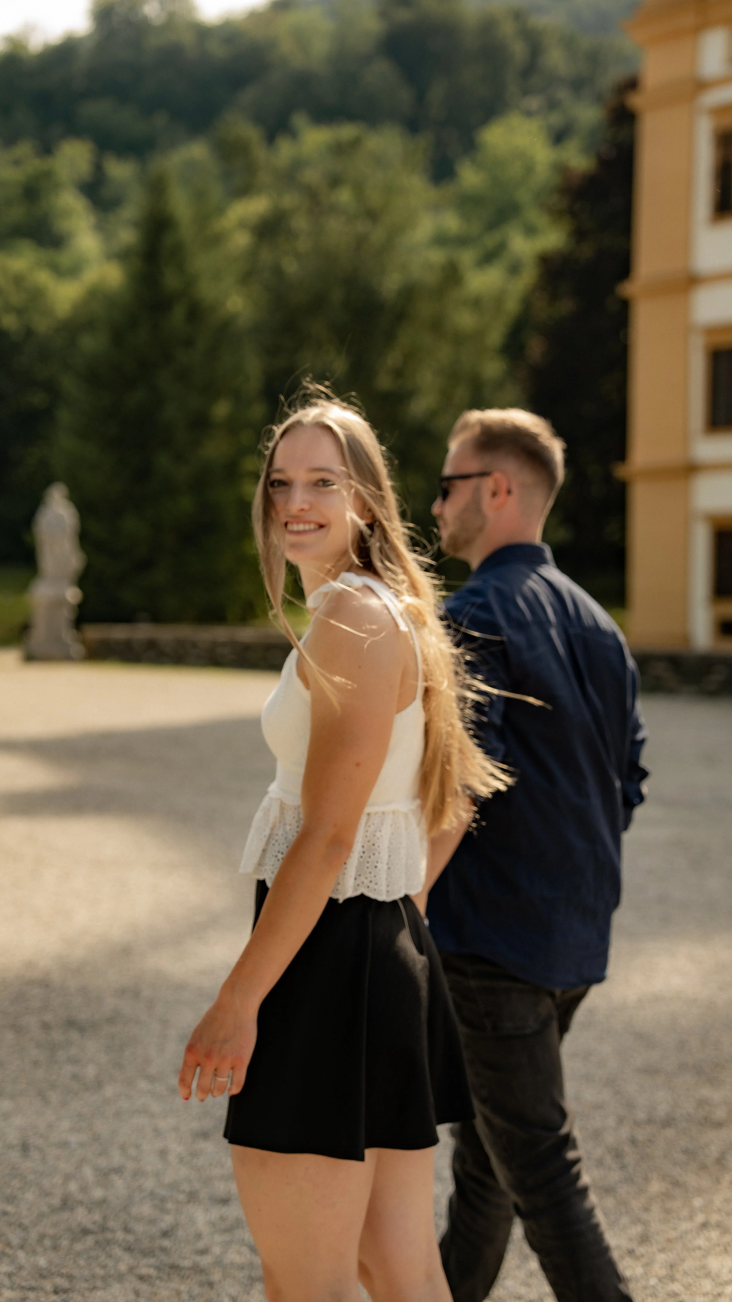 Ein lachendes Mädchen mit langem, blondem Haar trägt eine weiße Top und schwarze Shorts, während ein Mann mit Brille und schwarzem Jacke im Hintergrund steht, beide im Freien bei sonnigem Wetter.