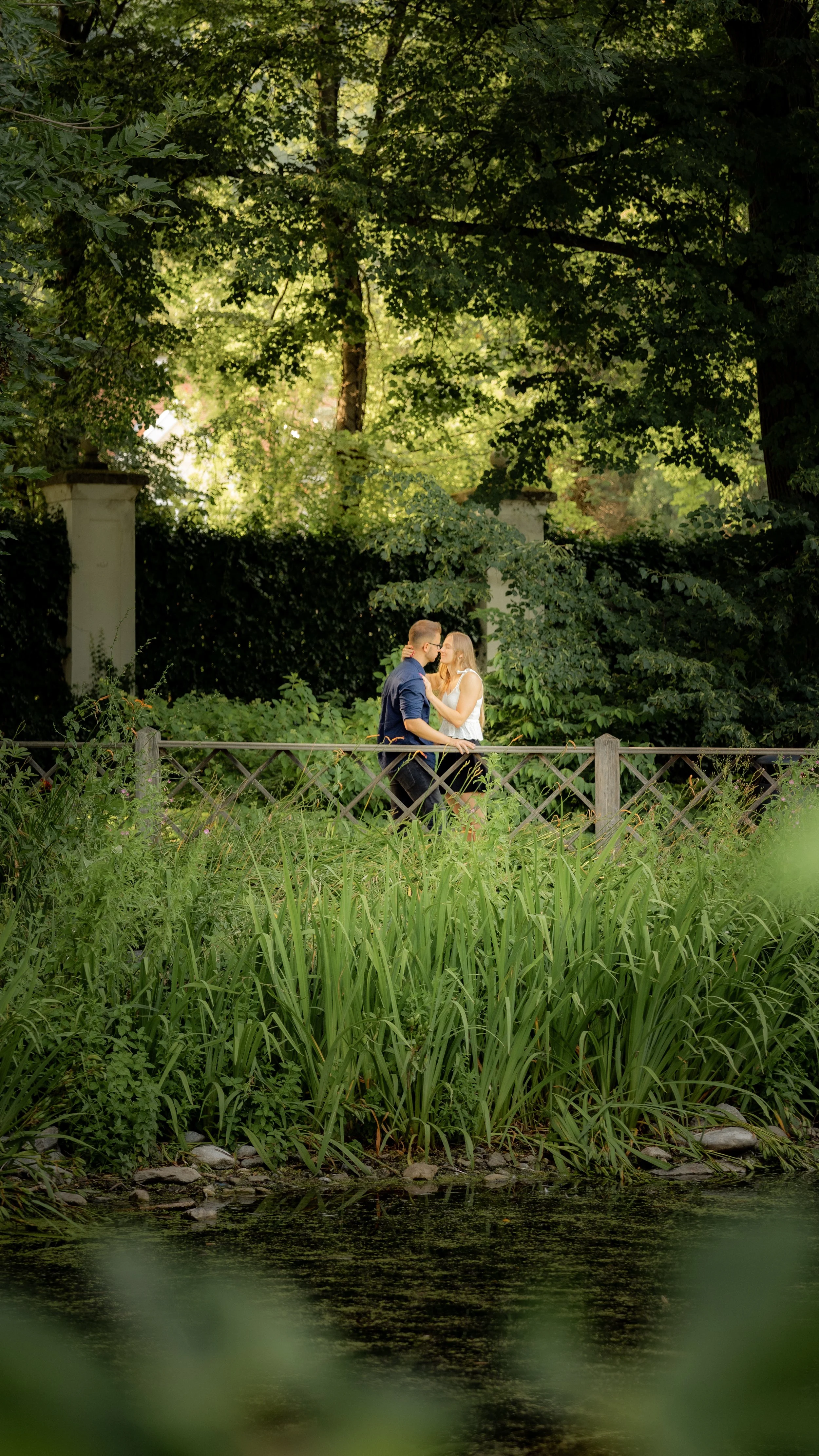 Ein junges Paar steht sich nahe am Zaun am Seeufer und küsst sich in einem grünen Park auf einer Bank.