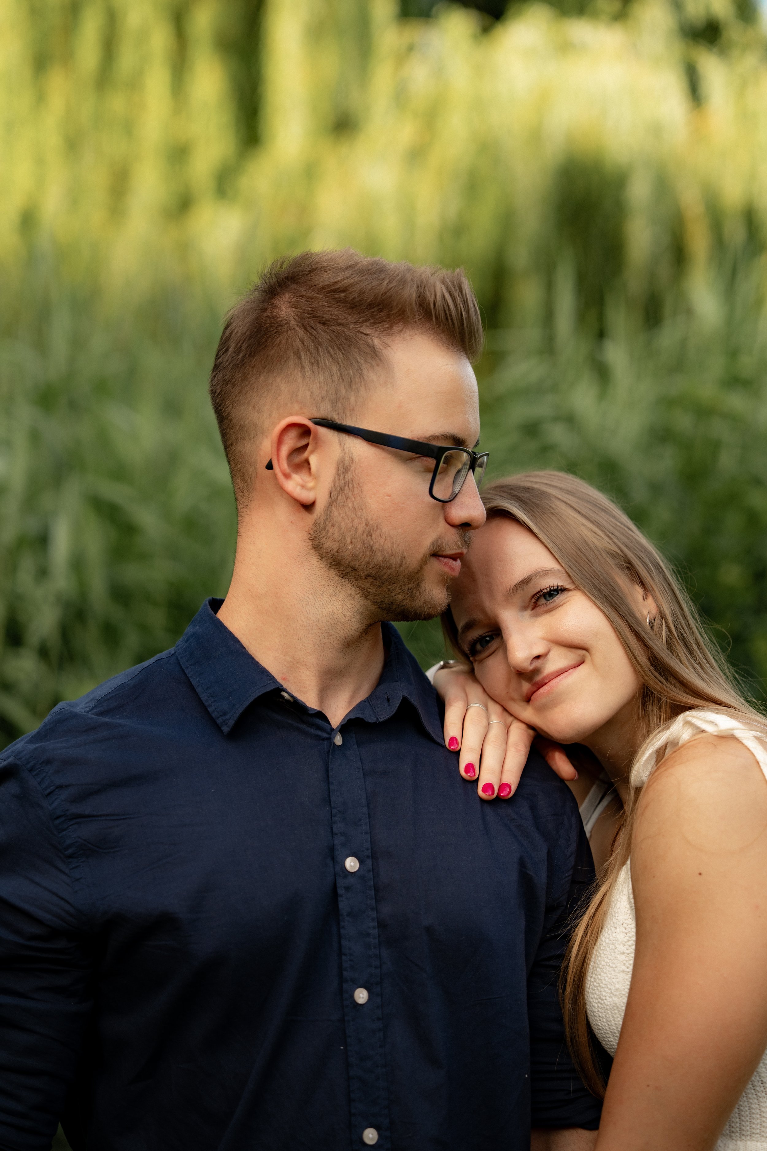Ein junges Paar steht im Grünen, sie lächelt, während er sie freundlich ansieht, umgeben von grünen Pflanzen.