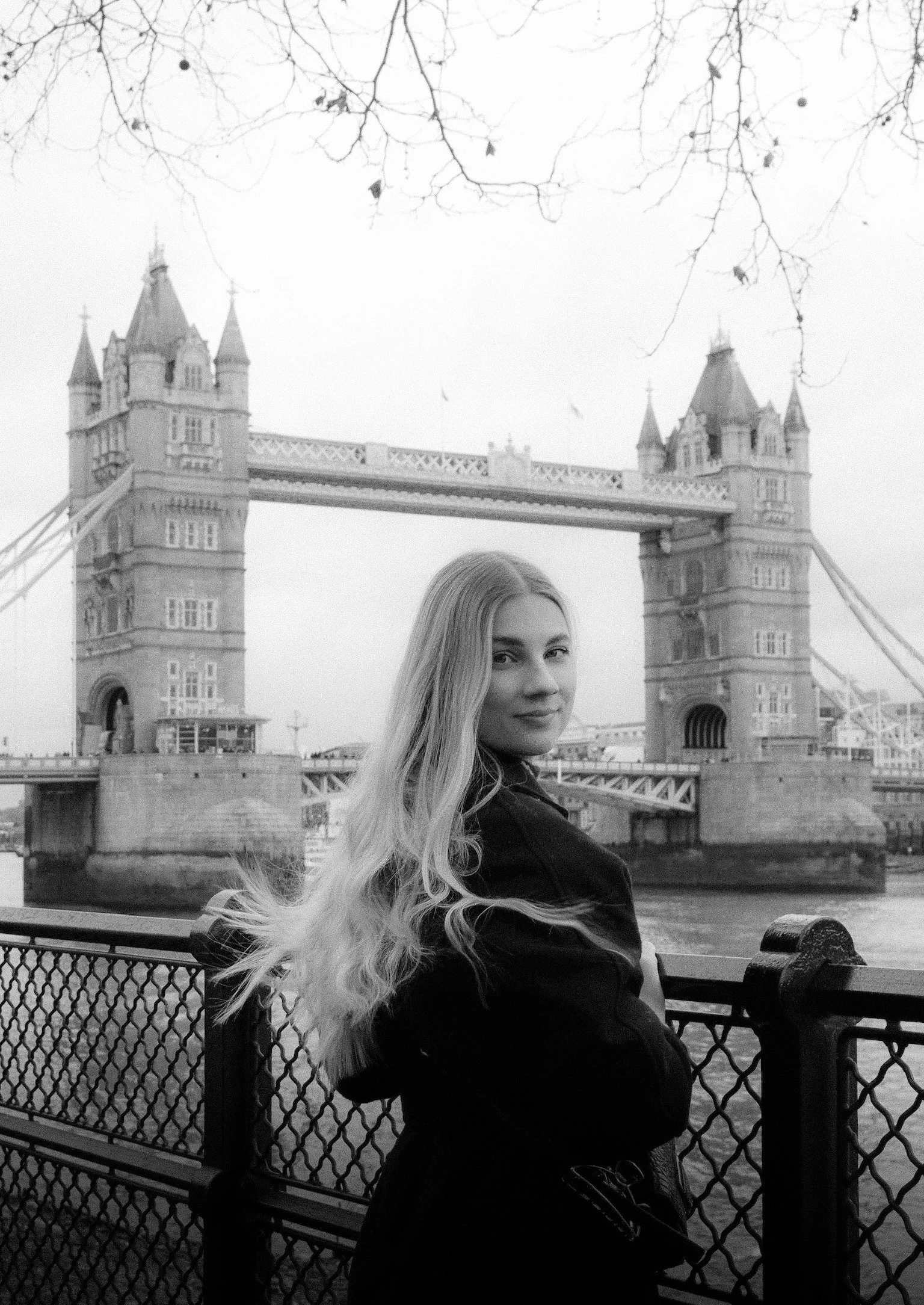 Eine junge Frau mit langen, hellen Haaren sitzt an einem Flussufer vor der Tower Bridge in London, bei bewölktem Himmel.