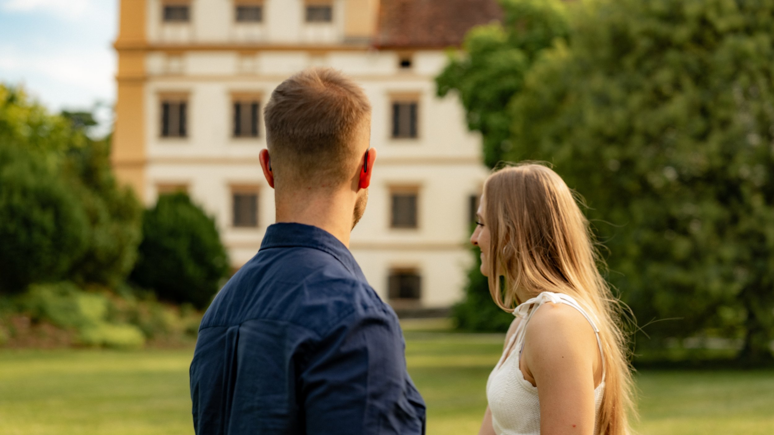 Ein Mann und eine Frau stehen sich im Park gegenüber, im Hintergrund ein mehrstöckiges Gebäude, umgeben von Bäumen, bei Sonnenuntergang.