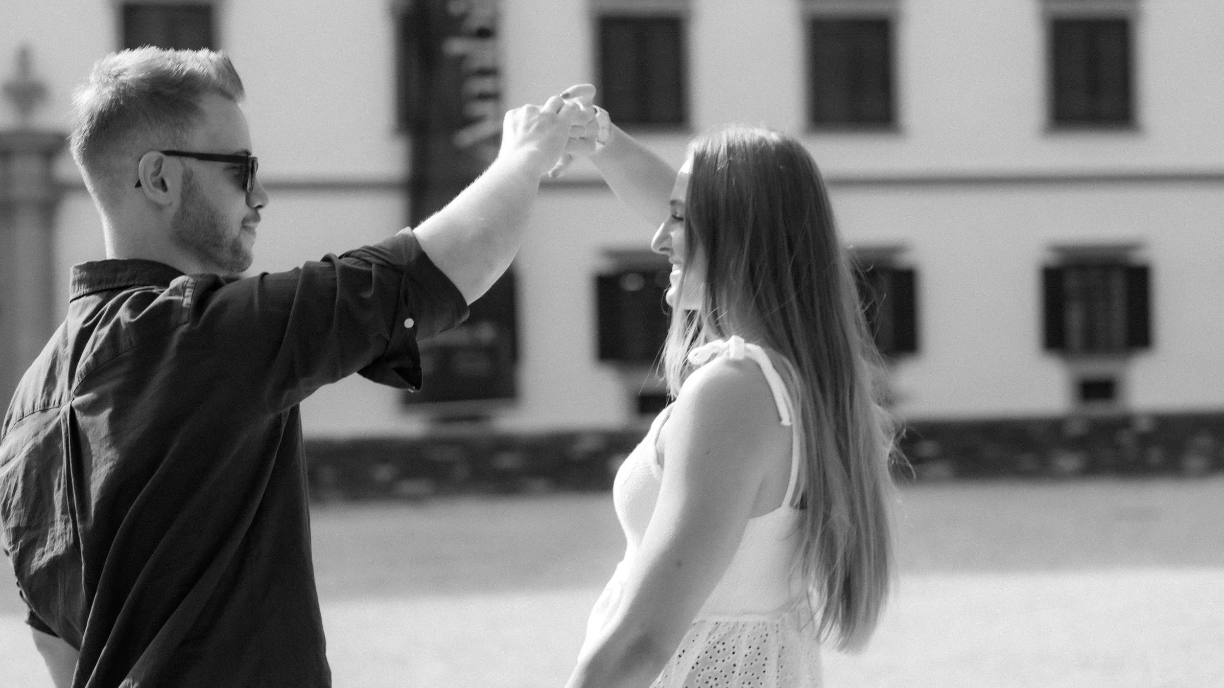 Ein junger Mann mit Sonnenbrille schneidet einer lachenden jungen Frau die Haare im Freien, im Hintergrund ein Gebäude.