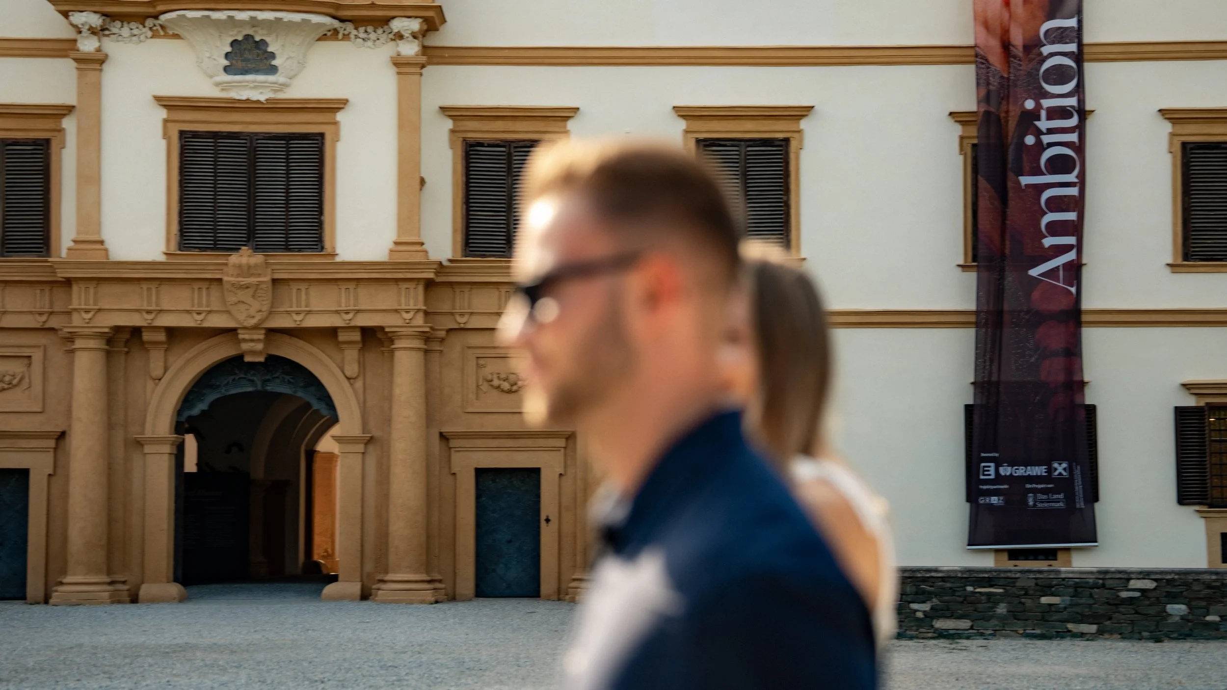 Menschen stehen vor einem historischen Gebäude, das mit einem großen schwarzen Banner mit weißem Text markiert ist.