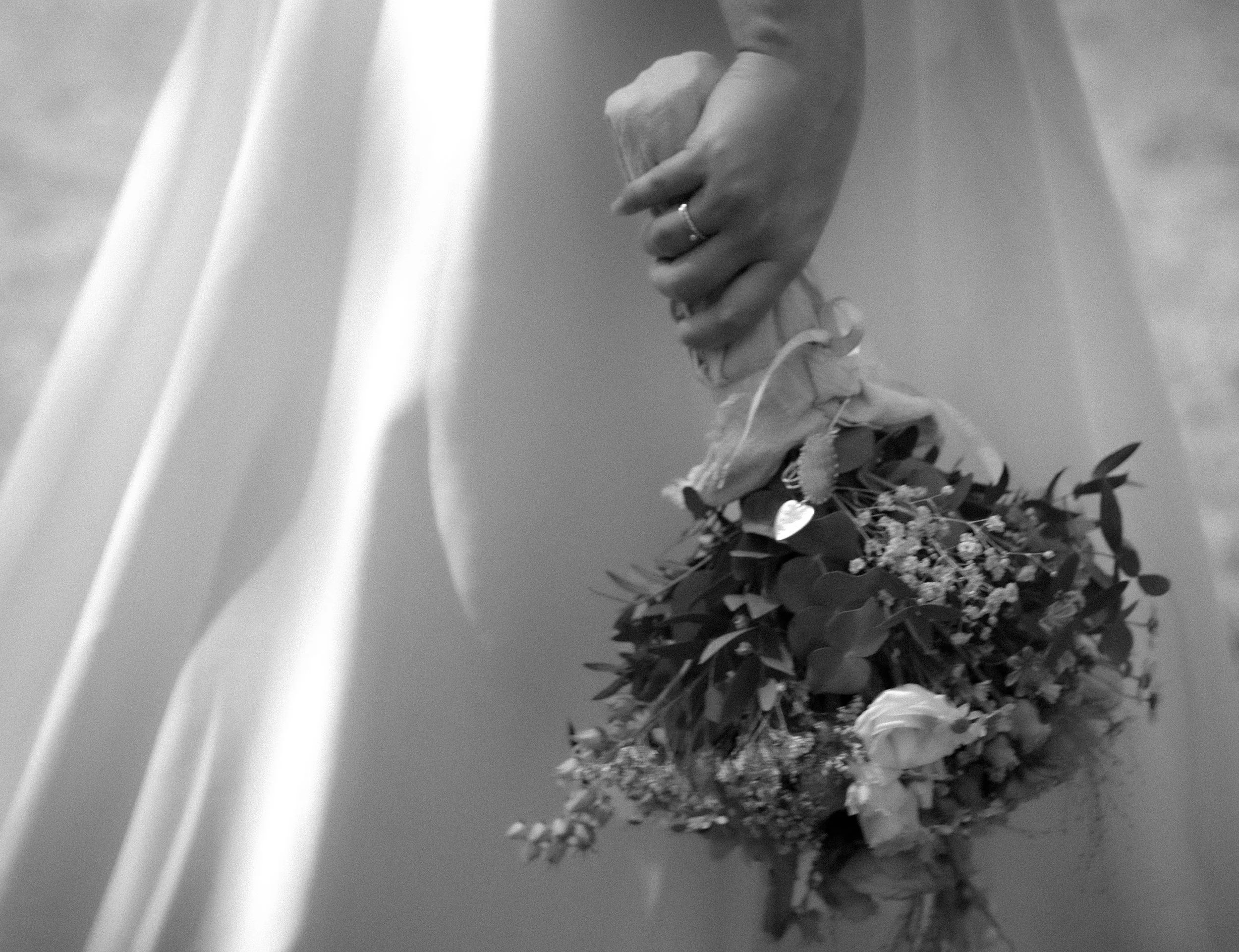 Schwarz-Weiß-Foto einer Hand, die eine Blumenstrauß hält, neben einem weißen Kleid und zarten Vorhängen im Hintergrund.
