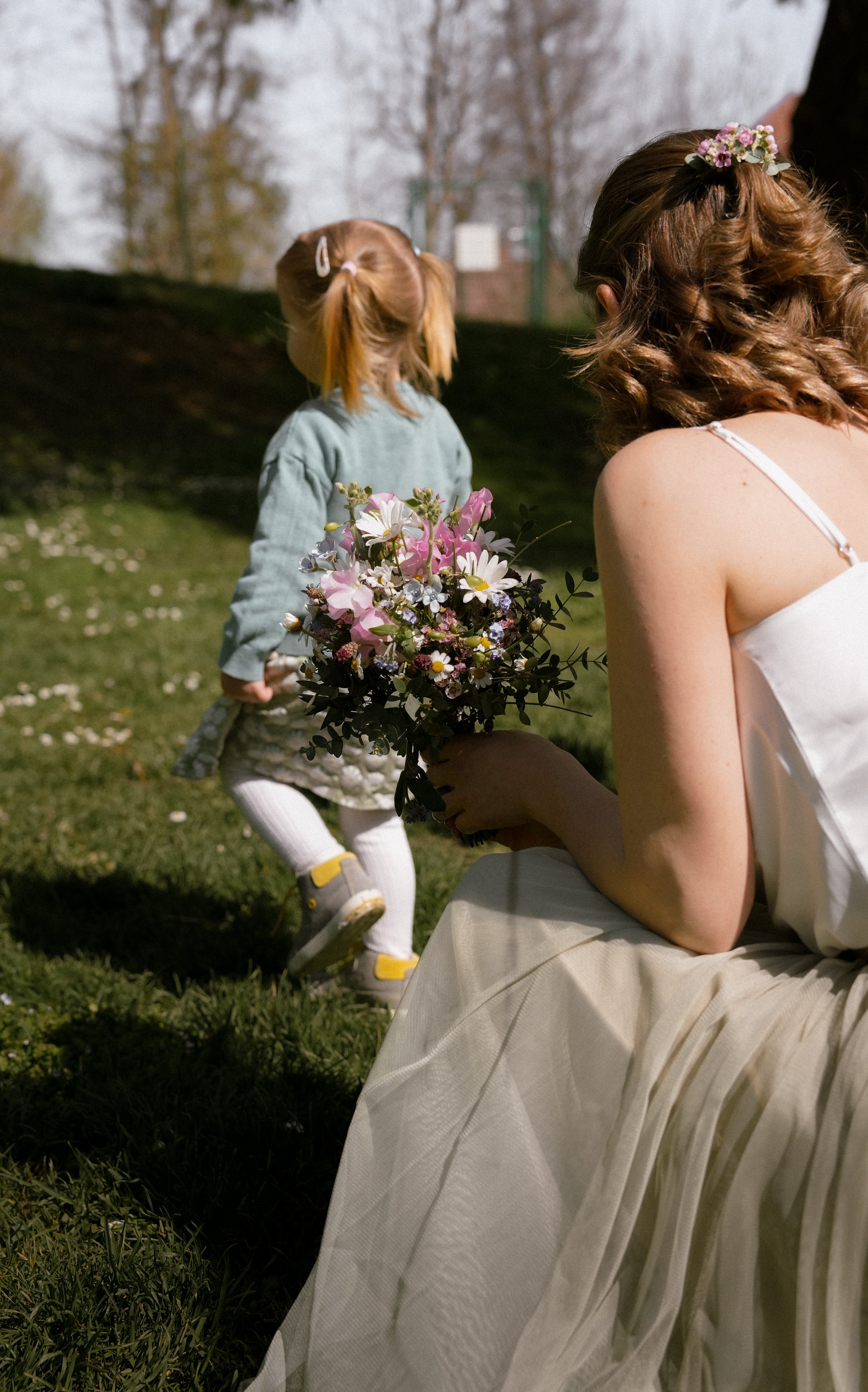 Eine Frau in weißem Kleid hält einen Blumenstrauß, während ein kleines Mädchen mit Zöpfen und grauem Kleid im Hintergrund in einem Park läuft.