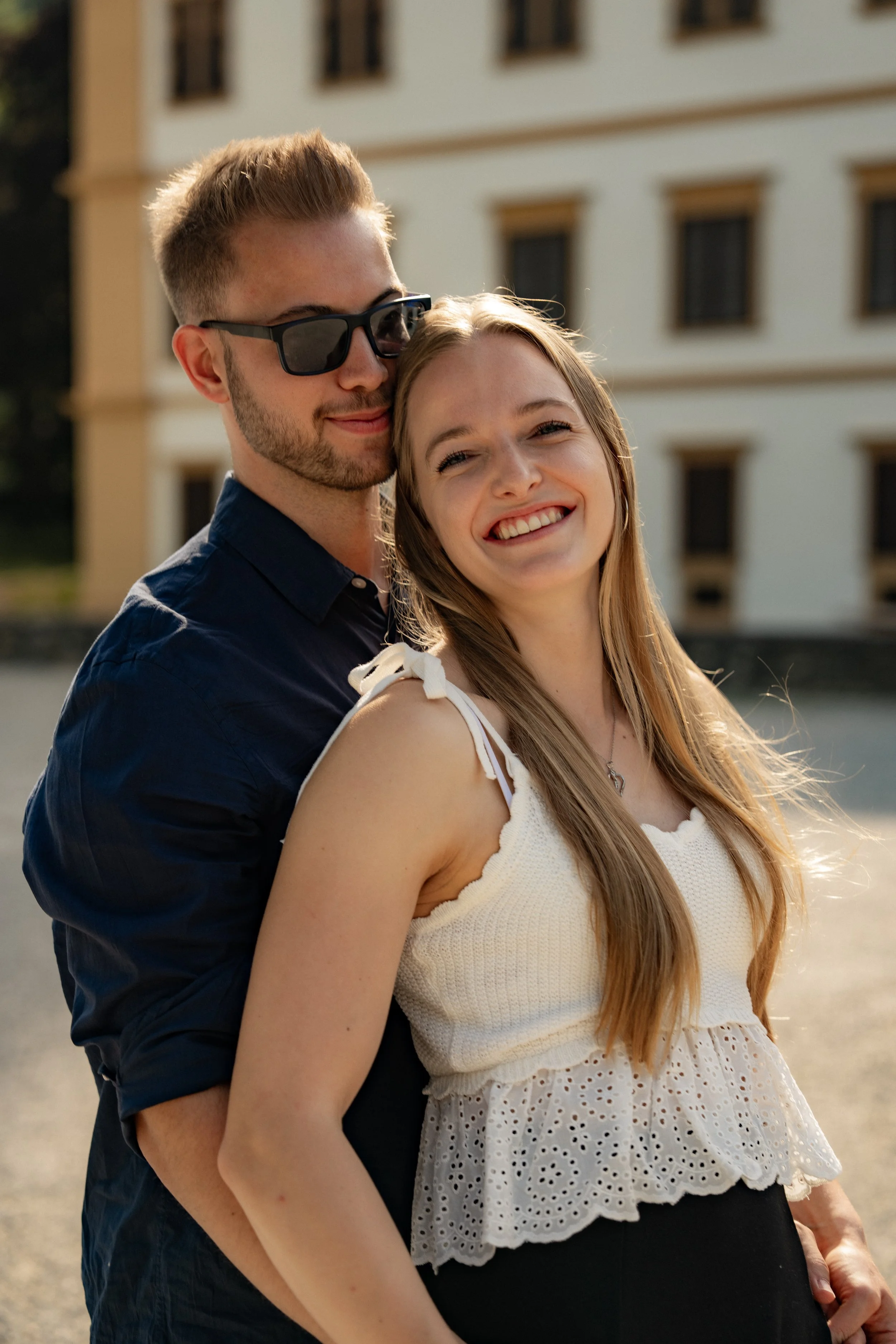 Ein lächelndes Paar, ein Mann mit dunklen Sonnenbrillen und eine Frau in einer weißen Top, steht draußen vor einem Gebäude, umgeben von Sonnenlicht.