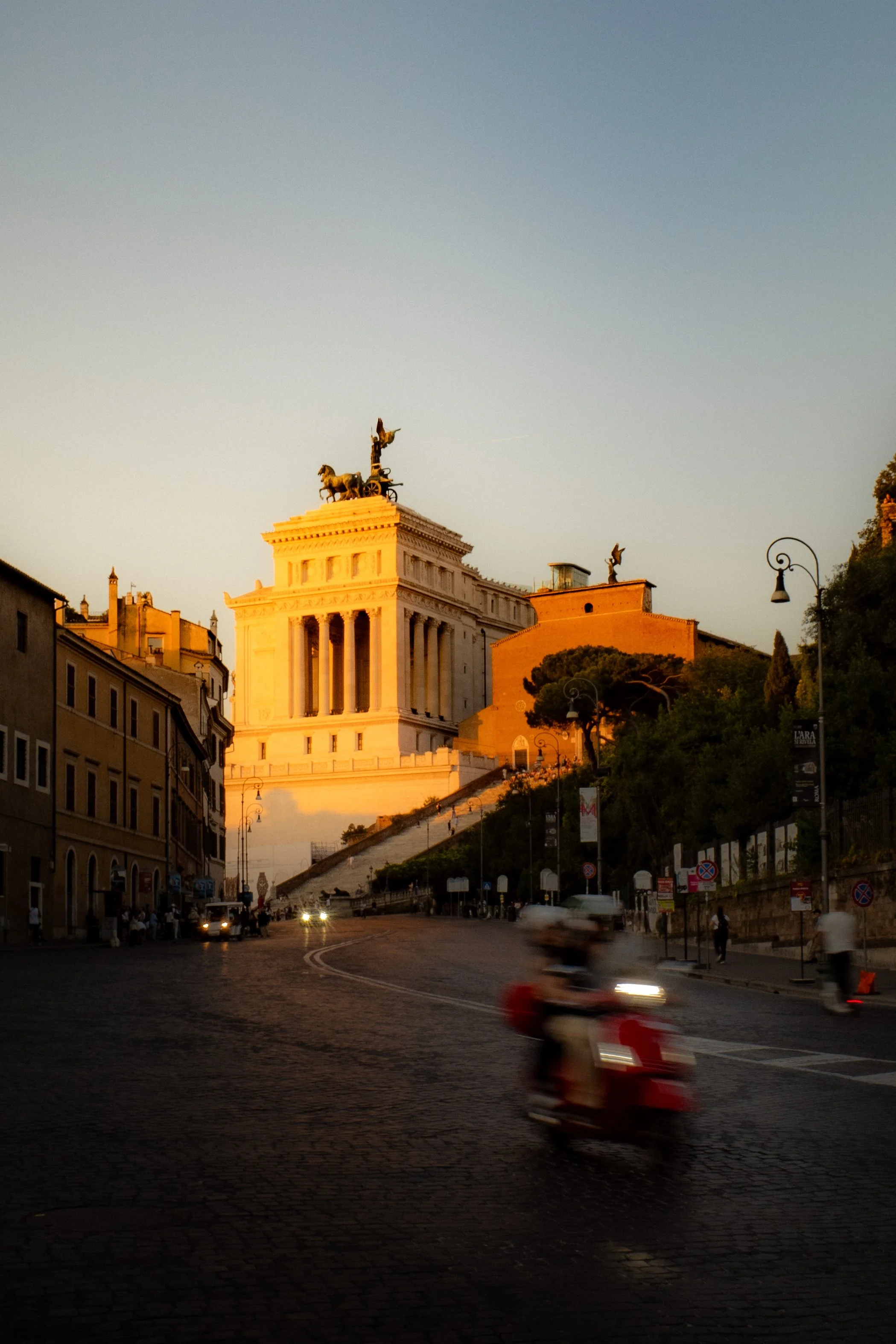 Das Bild zeigt eine Straße mit einigen fahrenden Autos und einem Motorroller im Vordergrund. Im Hintergrund ist das Victor-Emanuel-Denkmal auf der Piazza del Campidoglio in Rom bei Sonnenuntergang sichtbar, mit einem blauen Himmel und Gebäuden auf beiden Seiten der Straße.