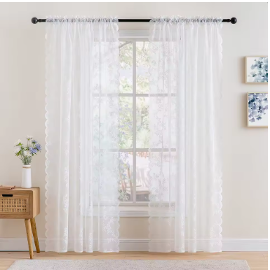White sheer curtains hanging in front of a window with trees visible outside, with a small plant on a wooden side table to the left and framed artwork on the wall to the right.
