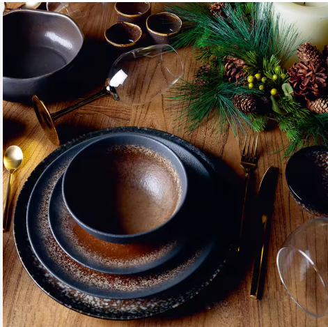 A festive table setting with black dinnerware, gold cutlery, a wine glass, and a holiday centerpiece with pinecones, green foliage, and berries.