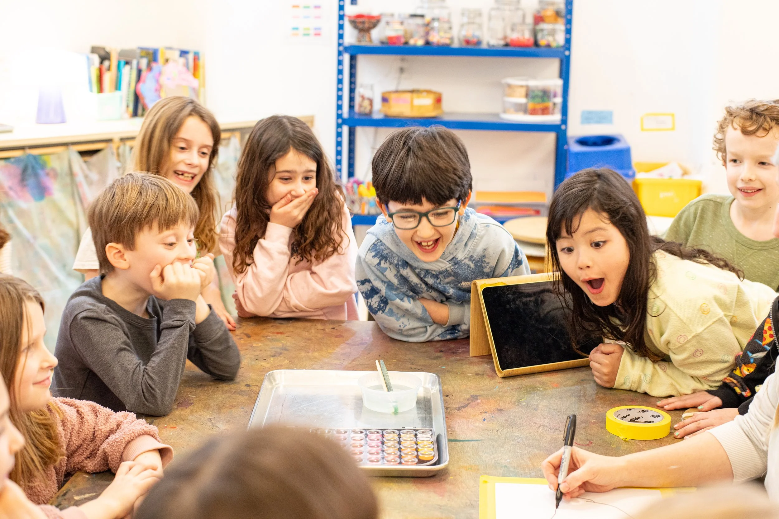 Children gathered around a table in a classroom, looking at a device with excited expressions.