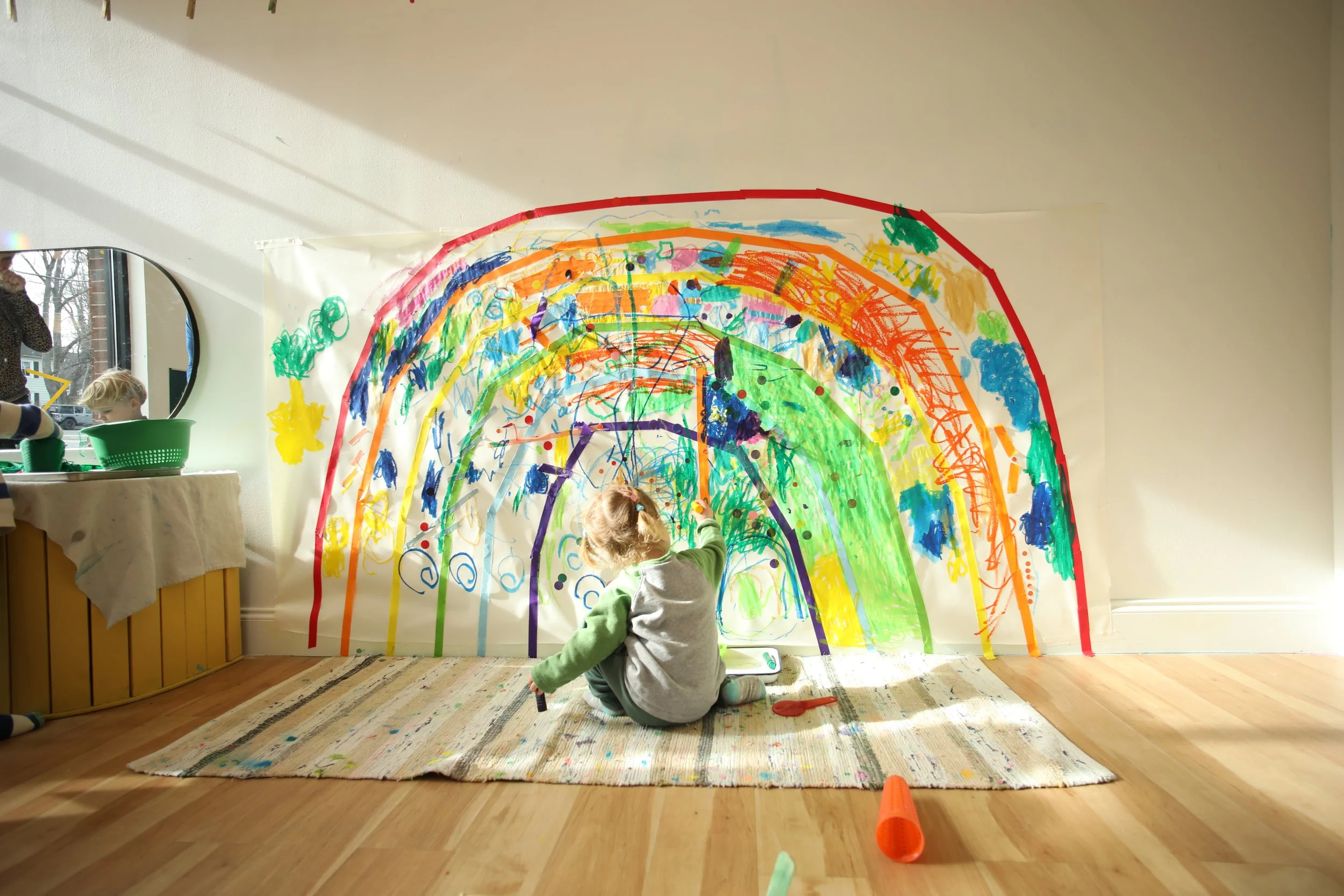 Child sitting on a rug painting a large rainbow mural on a wall with colorful markers and paints, with sunlight coming through a window.