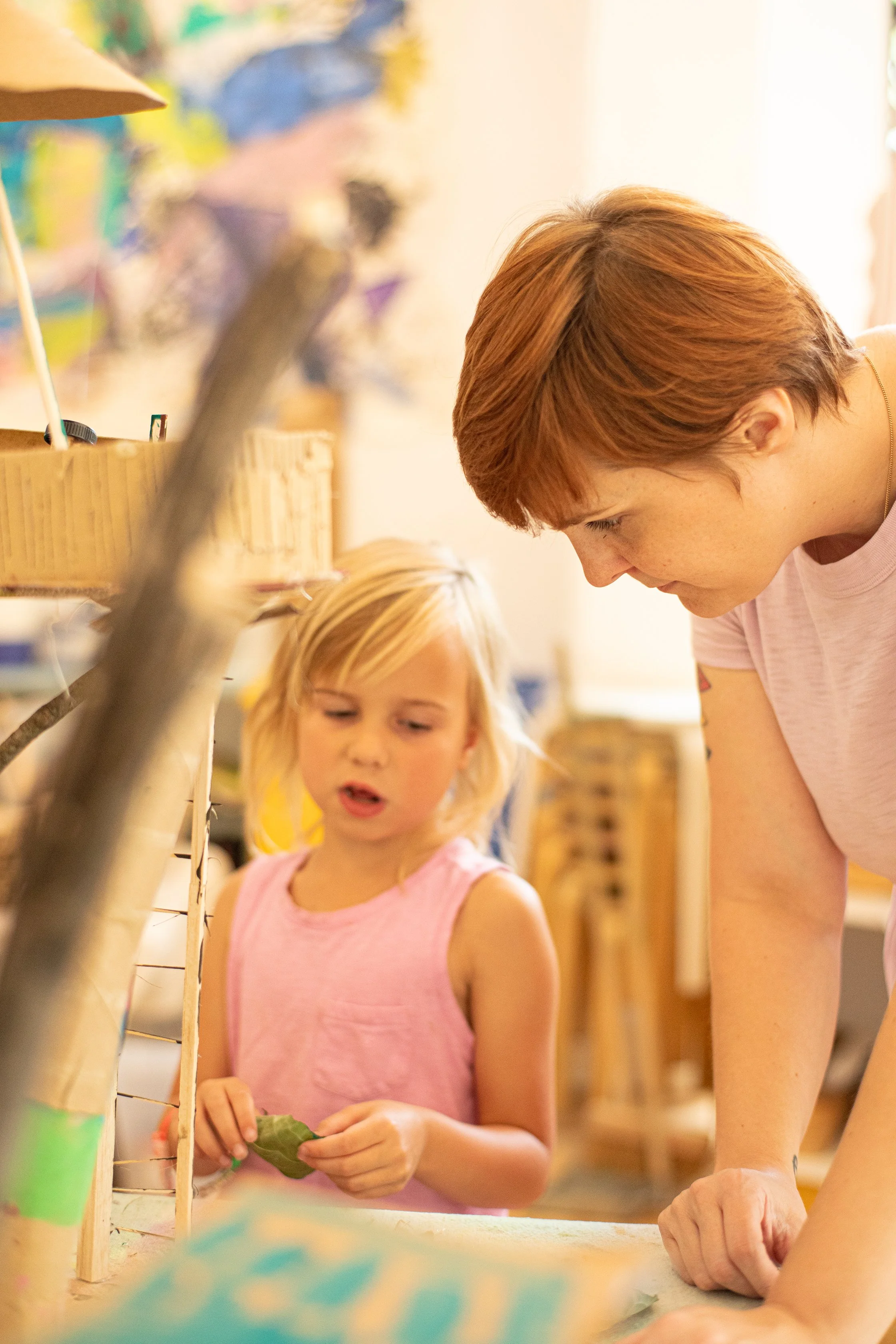 An adult woman and a young girl are working together on an art project in a colorful classroom. The woman is leaning over a table, and the girl is holding a leaf, looking at their project.