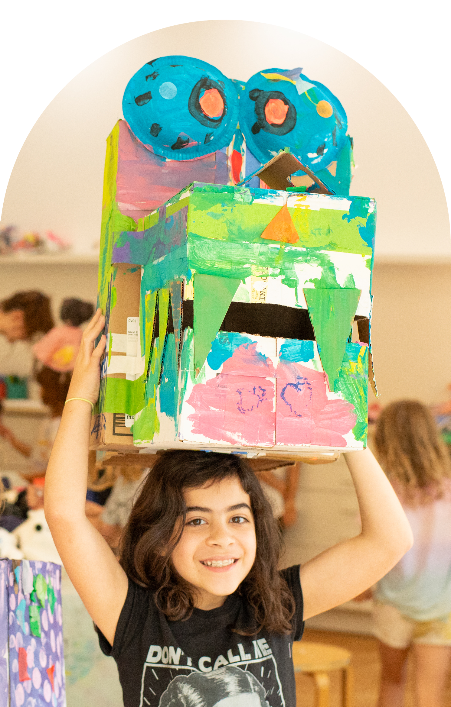 Girl holding a colorful cardboard sculpture of a robot with a box head and painted face at an art event.