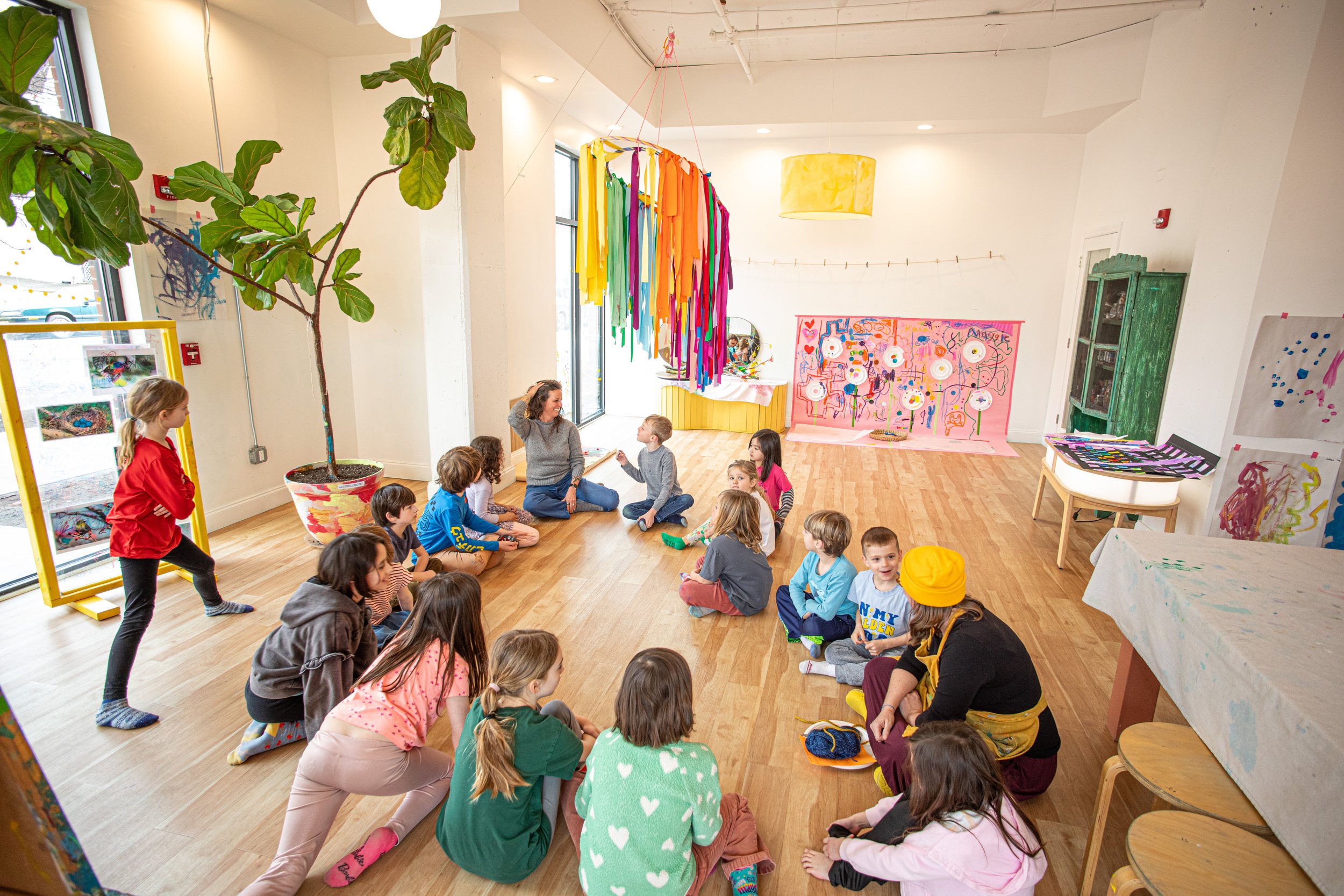 Children and a teacher sitting on the floor in a brightly decorated classroom during a storytelling or activity session.