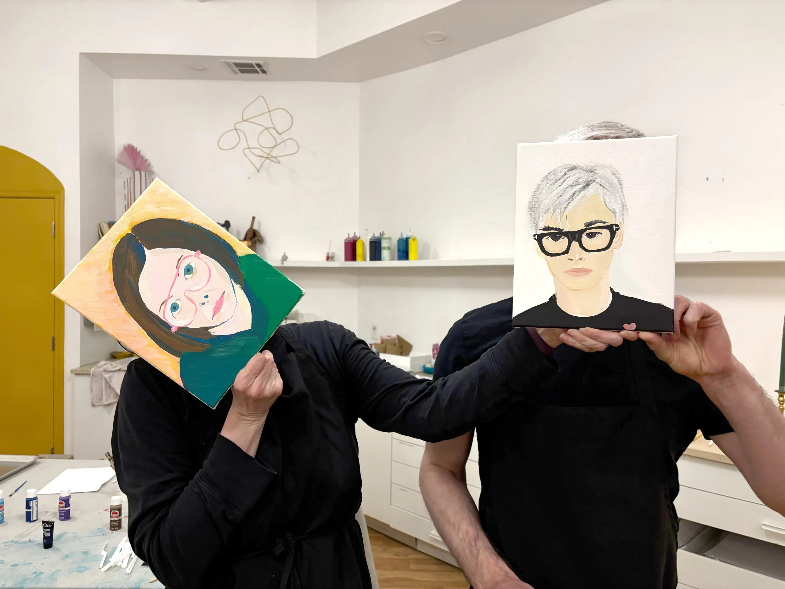 Two people are holding up paintings in a room with white walls. The person on the left is holding a colorful portrait of a woman with glasses, and the person on the right is holding a portrait of a person with short light hair and black glasses.
