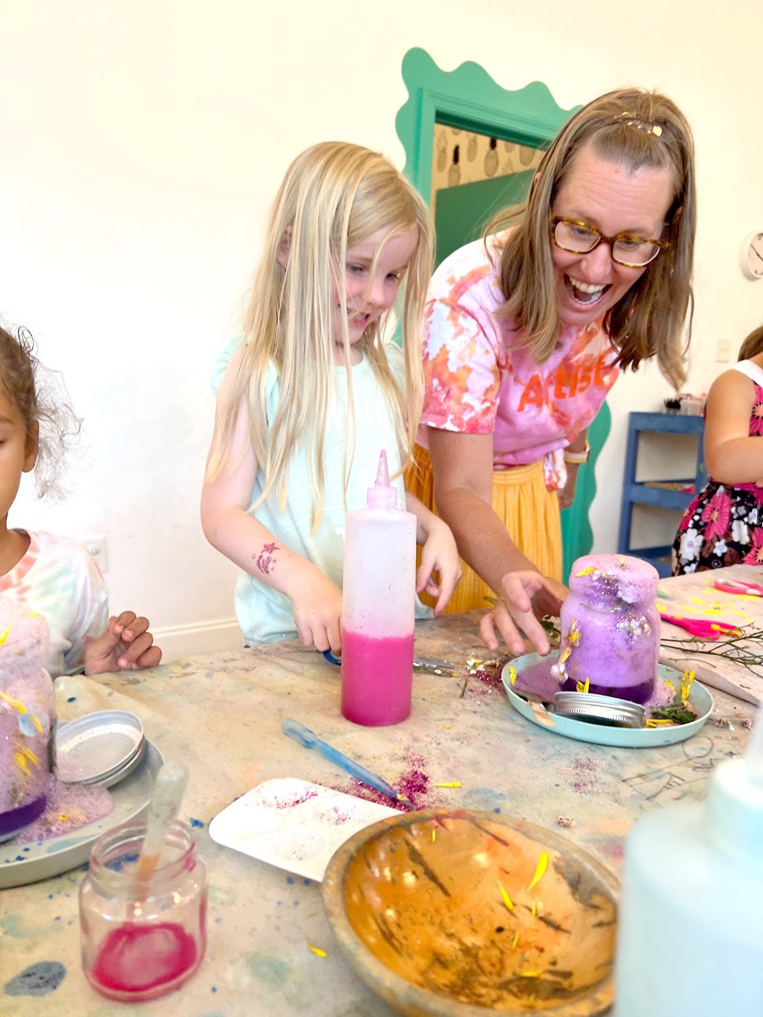 A group of children and an adult girl joyfully celebrating a birthday with decorated, colorful cakes and candles at a table.