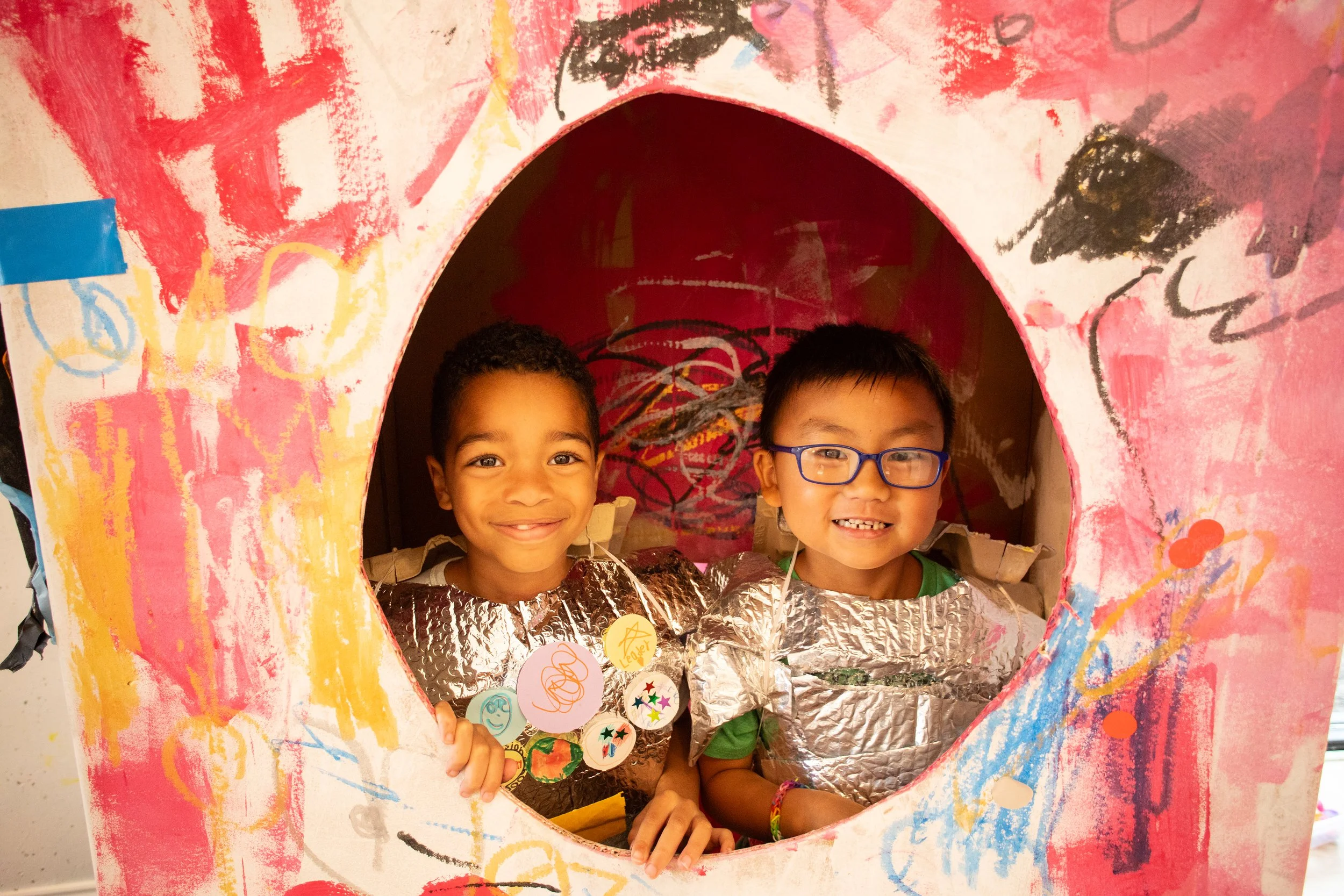 Two young boys smiling and posing behind a cardboard spaceship with colorful painted decorations, wearing shiny silver costumes and handmade badges.