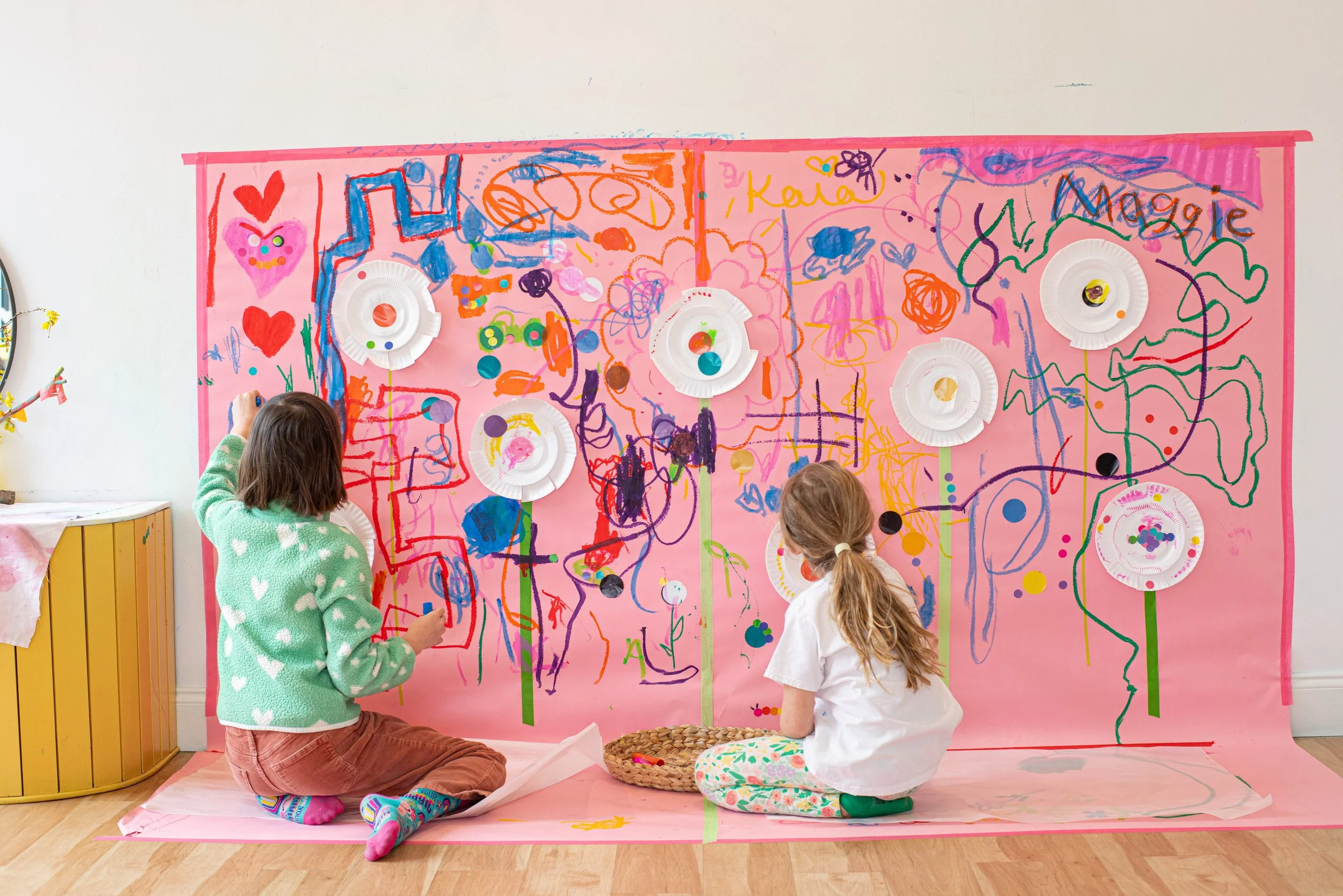 Two children, one with brown hair in a white shirt and green socks and the other with brown hair in a green sweater with white hearts, sit on the floor in front of a large pink paper backdrop decorated with colorful drawings and paper plates. The children are creating art with markers and stickers on the backdrop, which features scribbles, hearts, and flowers.